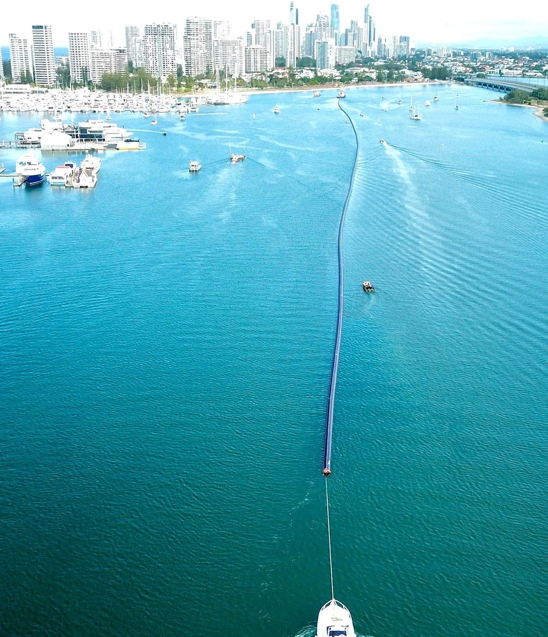 Aerial view of a marina and waterfront city with high-rise buildings, boats, and a long submerged floating pipeline with boats passing over it on the water.