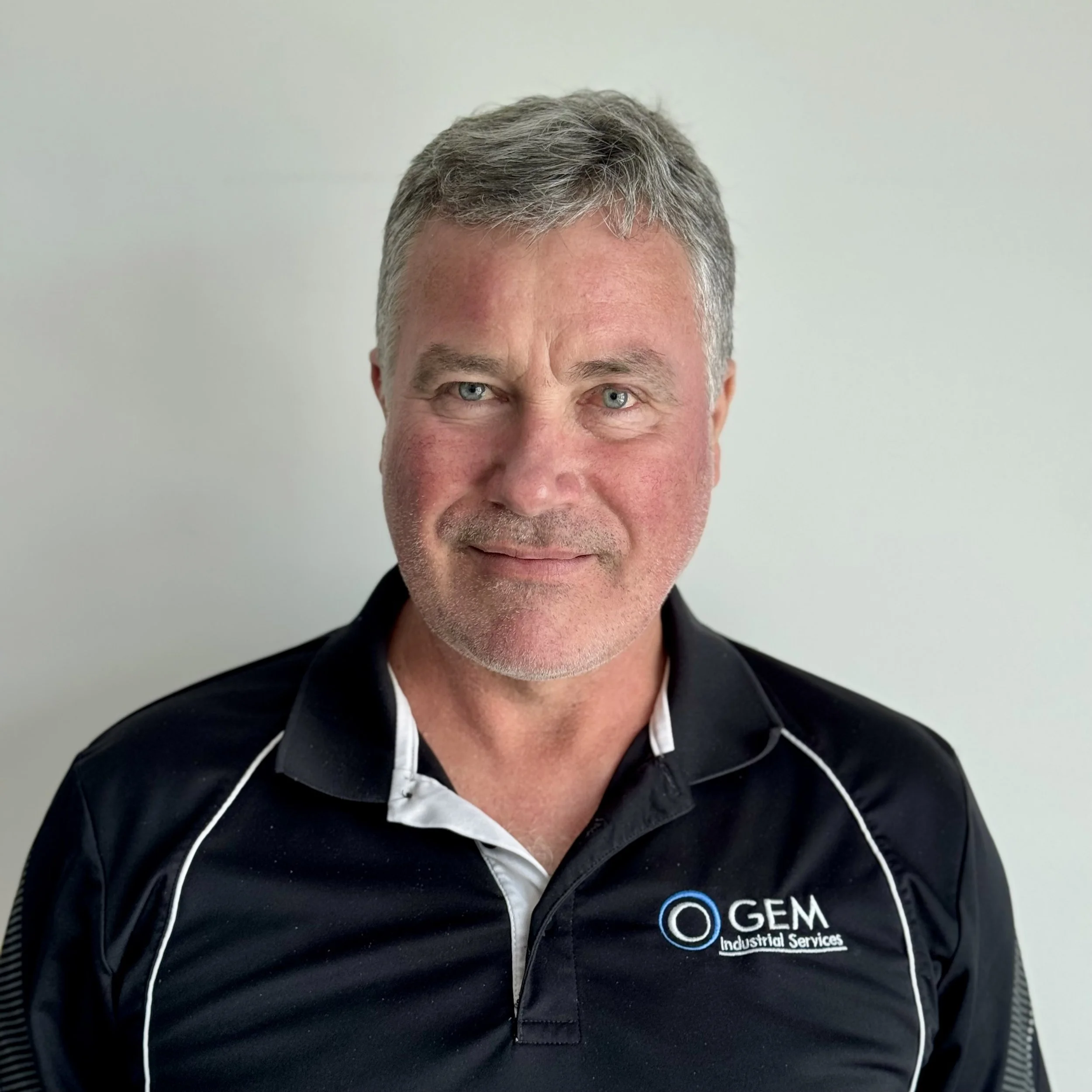 A middle-aged man with short gray hair, blue eyes, and a slight beard, wearing a black polo shirt with white accents and the logo of GEM Industrial Services.