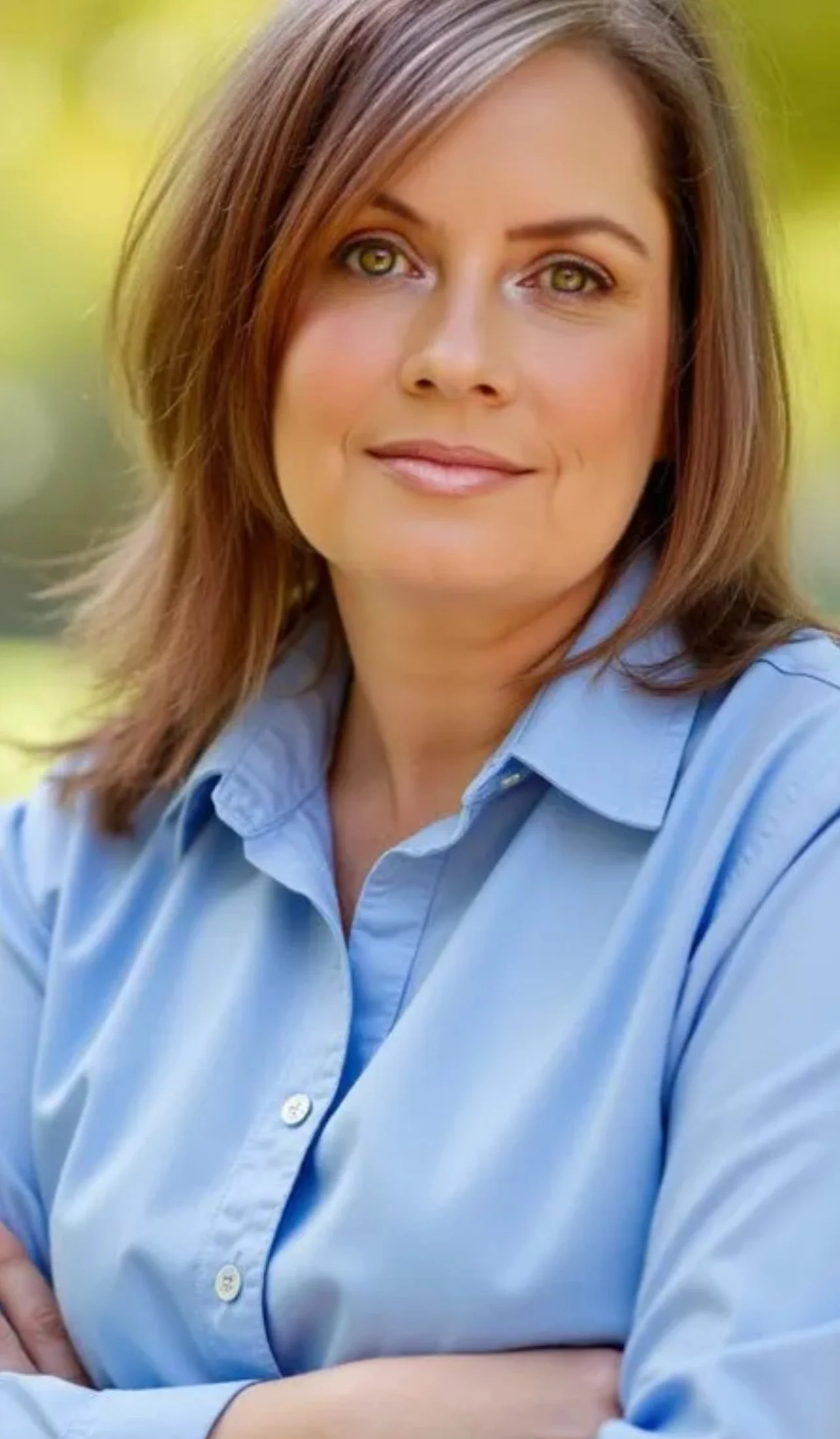 Colleen Murphy, Atkinson, NH, a woman with shoulder-length auburn hair, light skin, and green eyes, smiling, wearing a light blue button-up shirt, standing outdoors with blurred green foliage in the background.