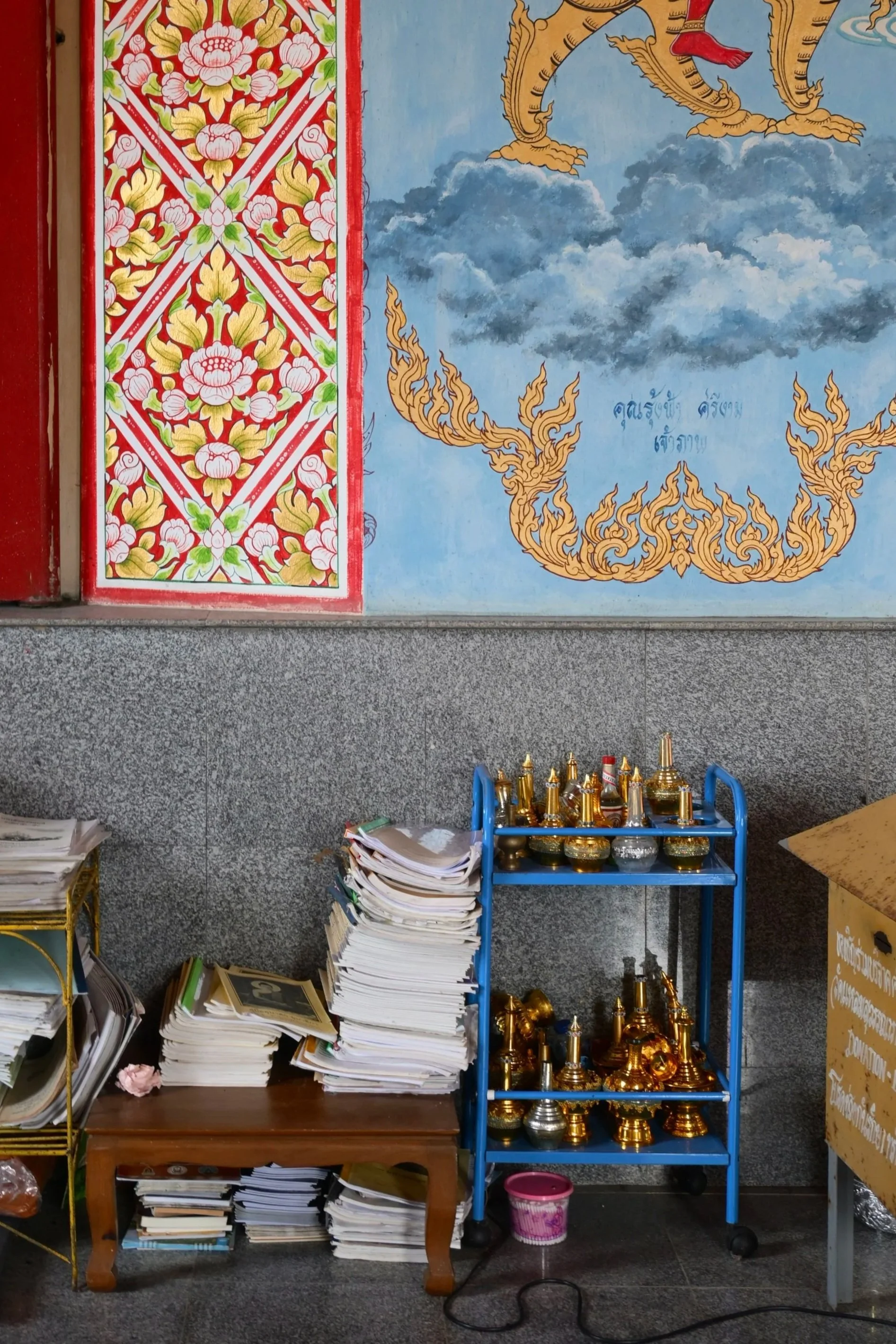 Stacked papers and small shelves with golden and silver religious artifacts in front of a decorated wall with Thai art and script.
