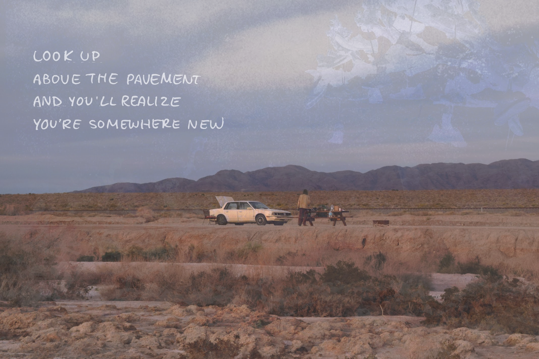 A desert landscape with mountains in the background. There is a person standing near a vintage car. A picnic table and some supplies are also visible.