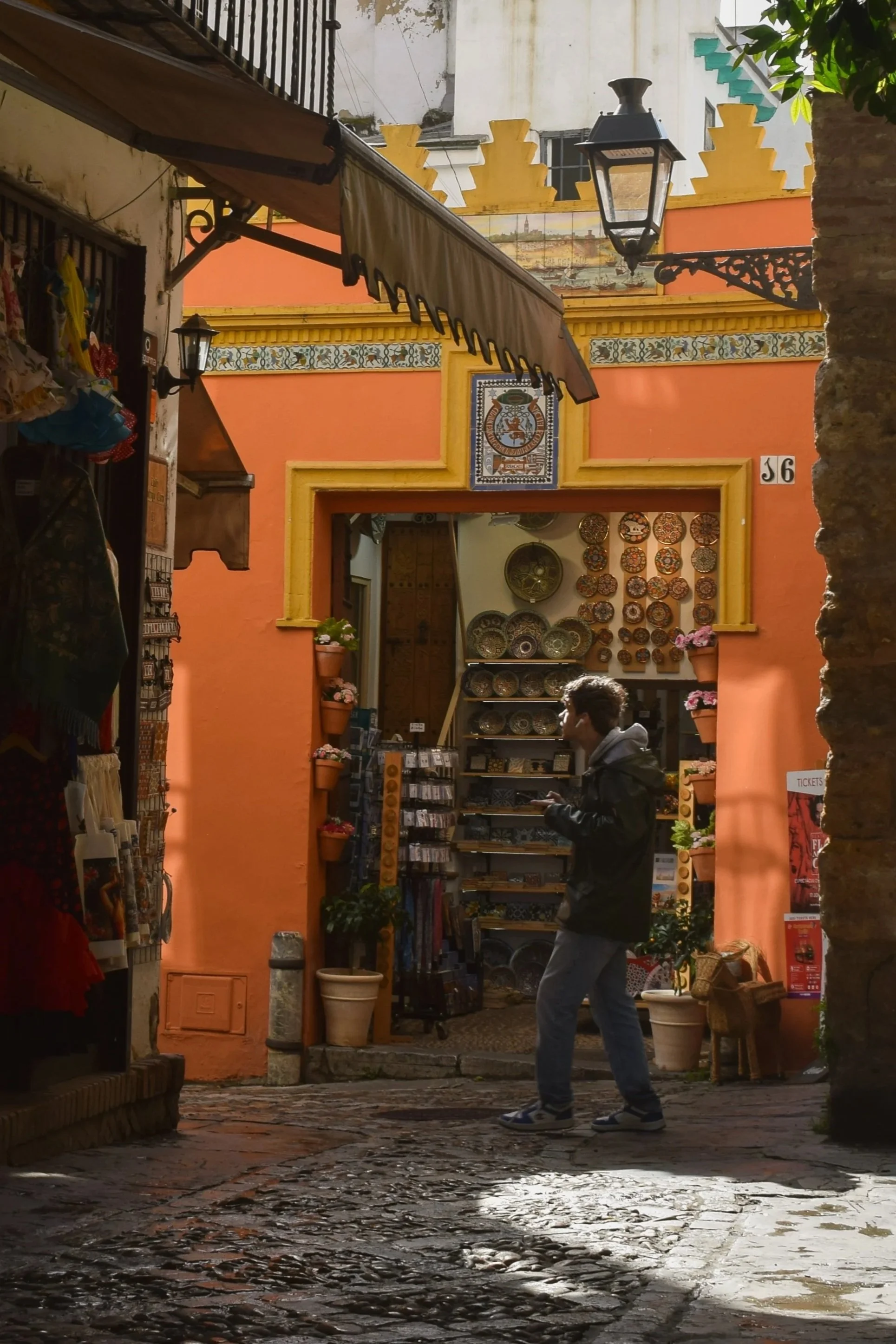 A person walking through a narrow, cobblestone street with colorful buildings. The entrance to a shop with plates and souvenirs is visible, with potted plants and a decorative sign above the doorway.