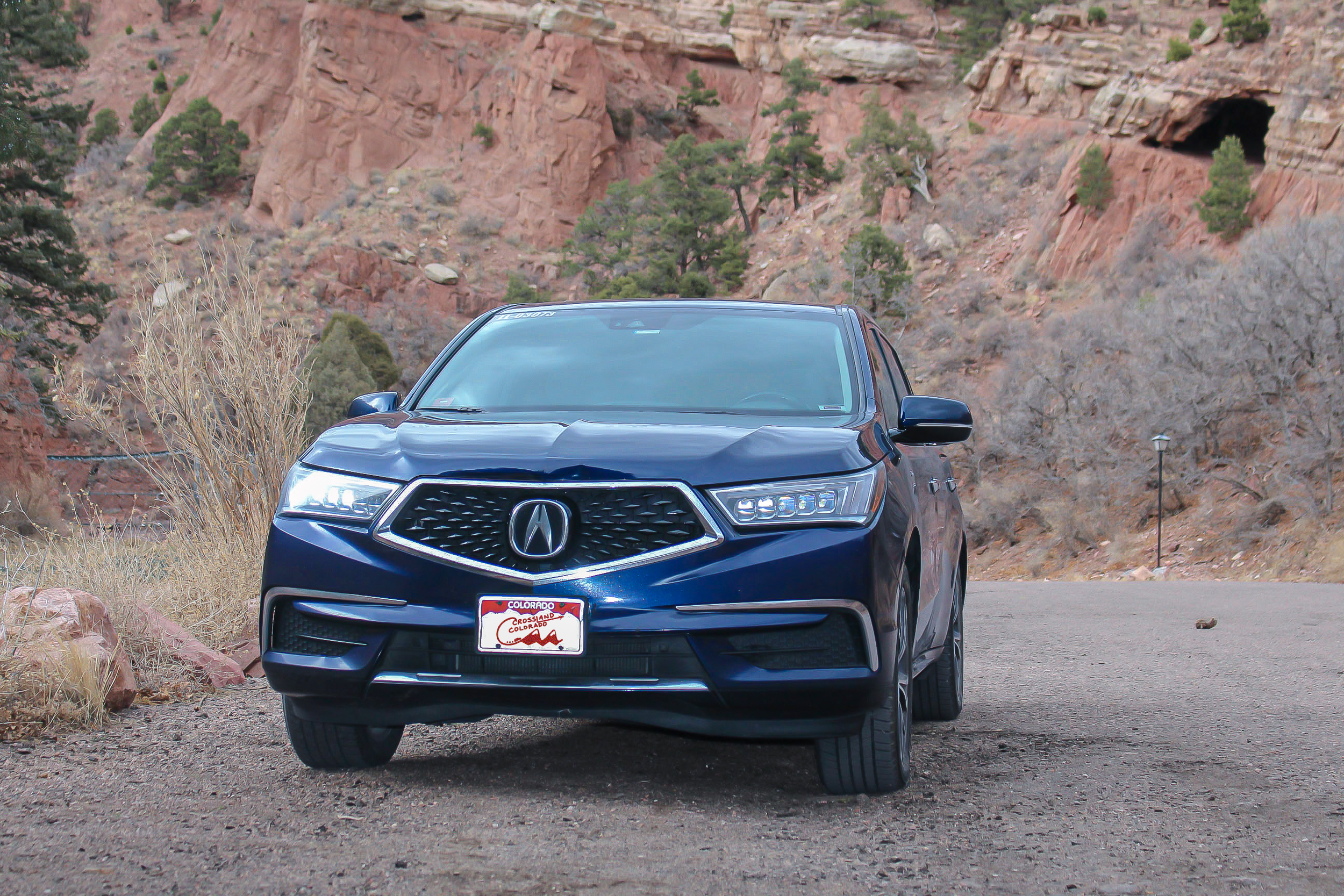 A blue Acura SUV parked on a dirt road in a mountainous area with reddish rock formations and sparse vegetation.