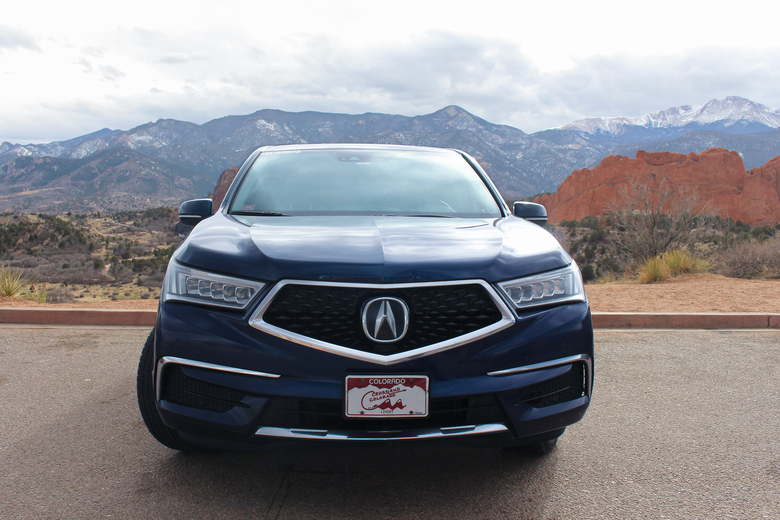 Front view of a dark blue Acura SUV with a Colorado license plate cover with Crossland Colorado on it, parked on a road with a mountainous landscape and red rock formations in the background.