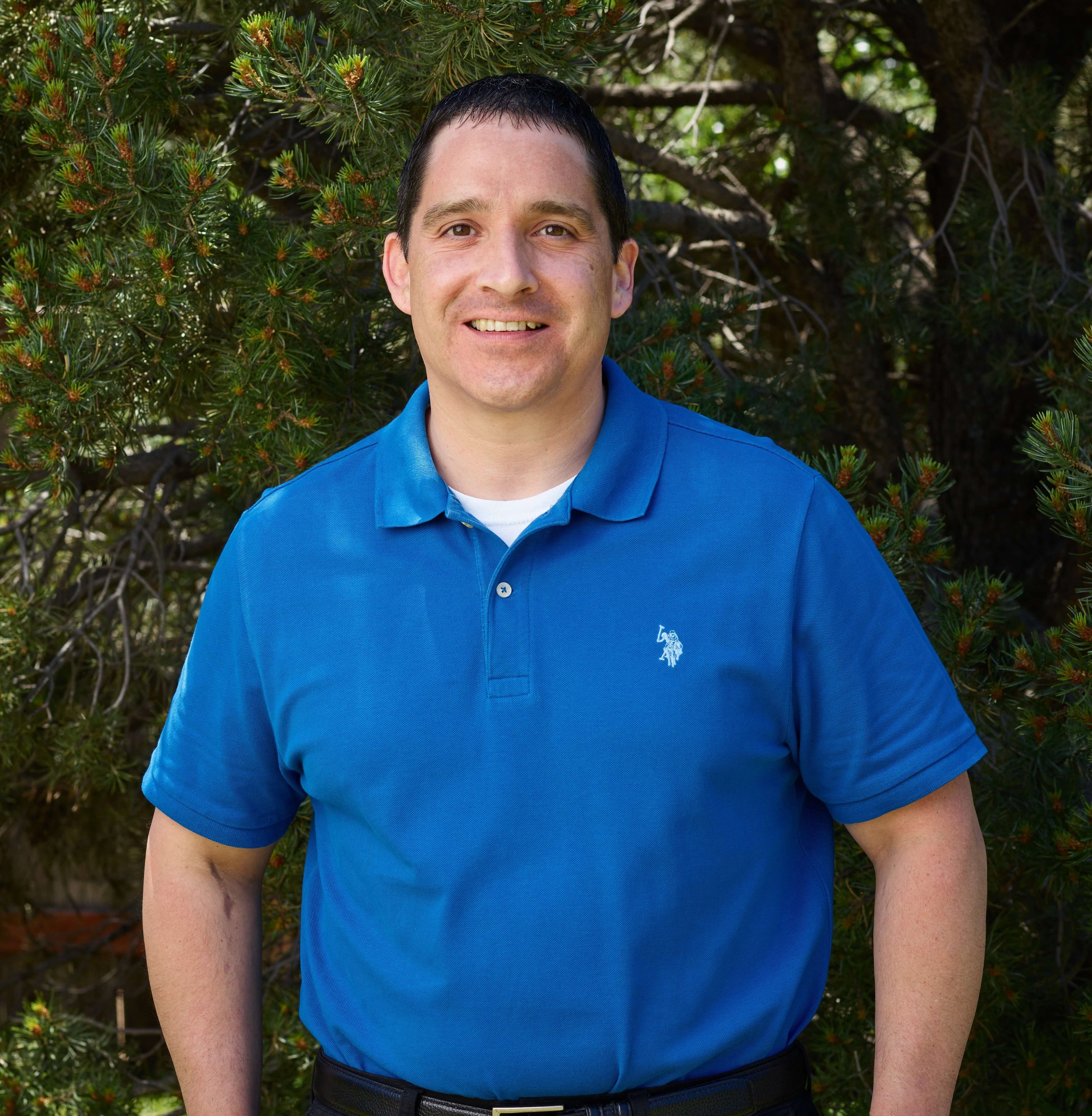 The owner, Marcus Manuel, standing outdoors in front of green pine tree, smiling, wearing a bright blue polo shirt.