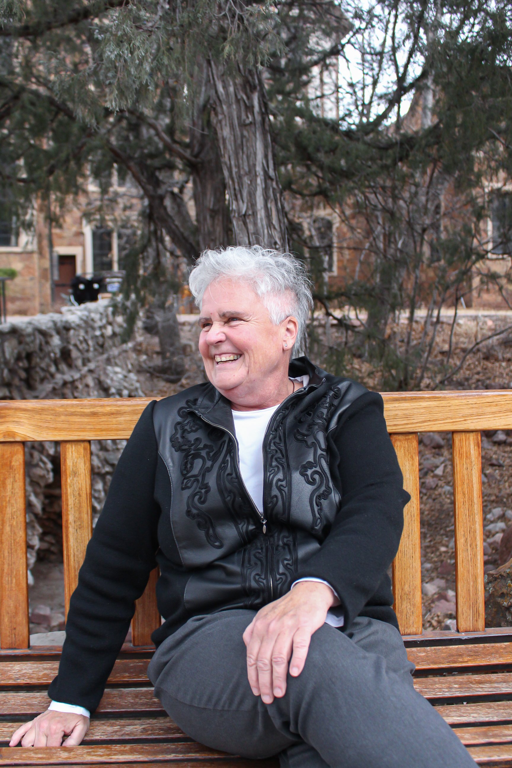 A smiling elderly person with gray curly hair sitting on a wooden bench outdoors, with a large tree and residential buildings in the background.