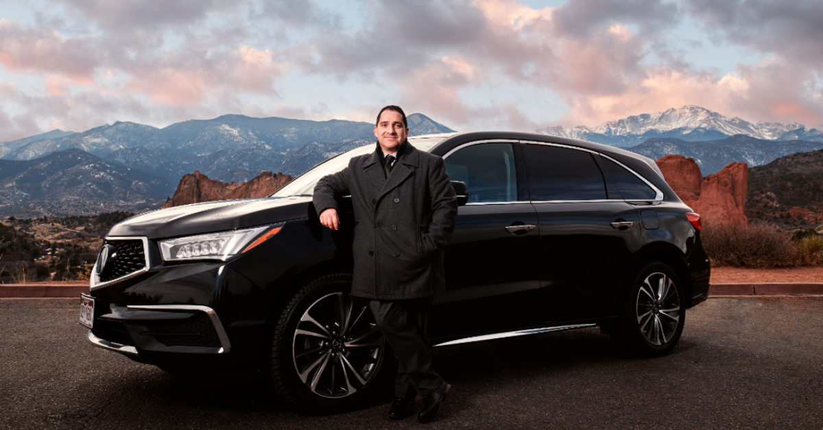 The owner, Marcus Manuel, in a dark coat leaning against a black luxury SUV parked on a scenic mountain road with snow-capped mountains and a partly cloudy sky in the background.