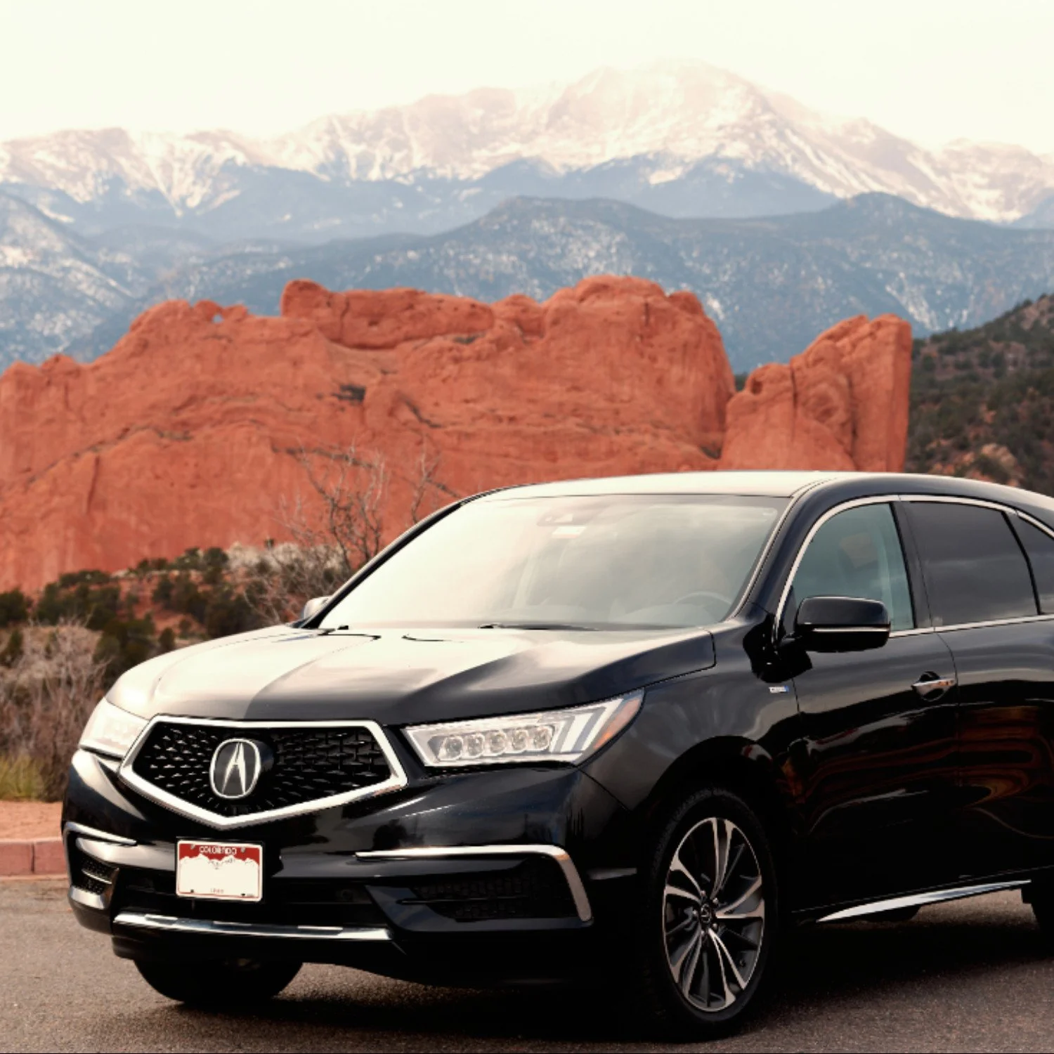 Black Acura SUV parked in front of red rock formations and snow-capped mountains during daytime.