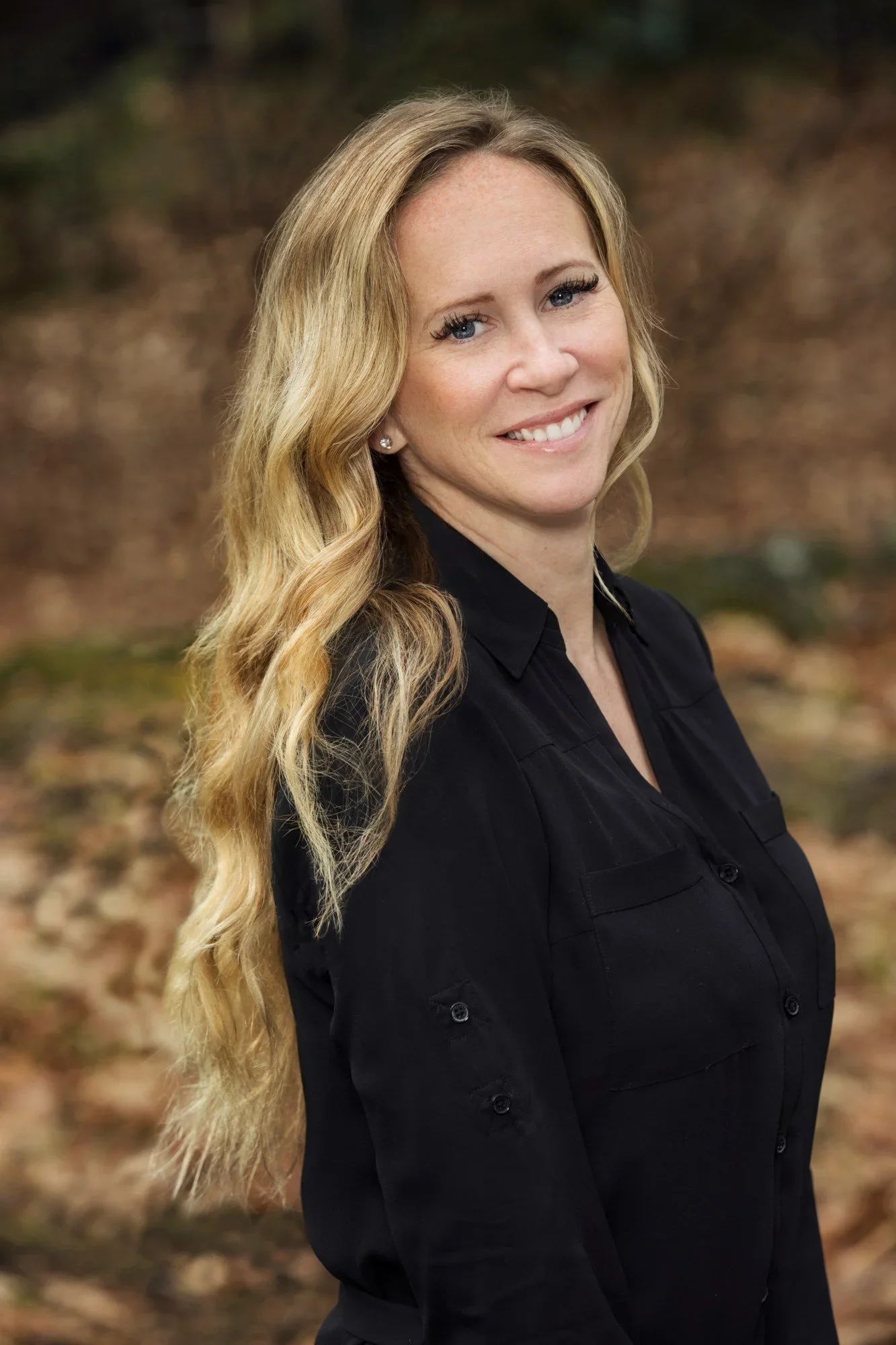 A woman with long, wavy blonde hair smiling outdoors, wearing a black button-up shirt, with a blurred natural background.