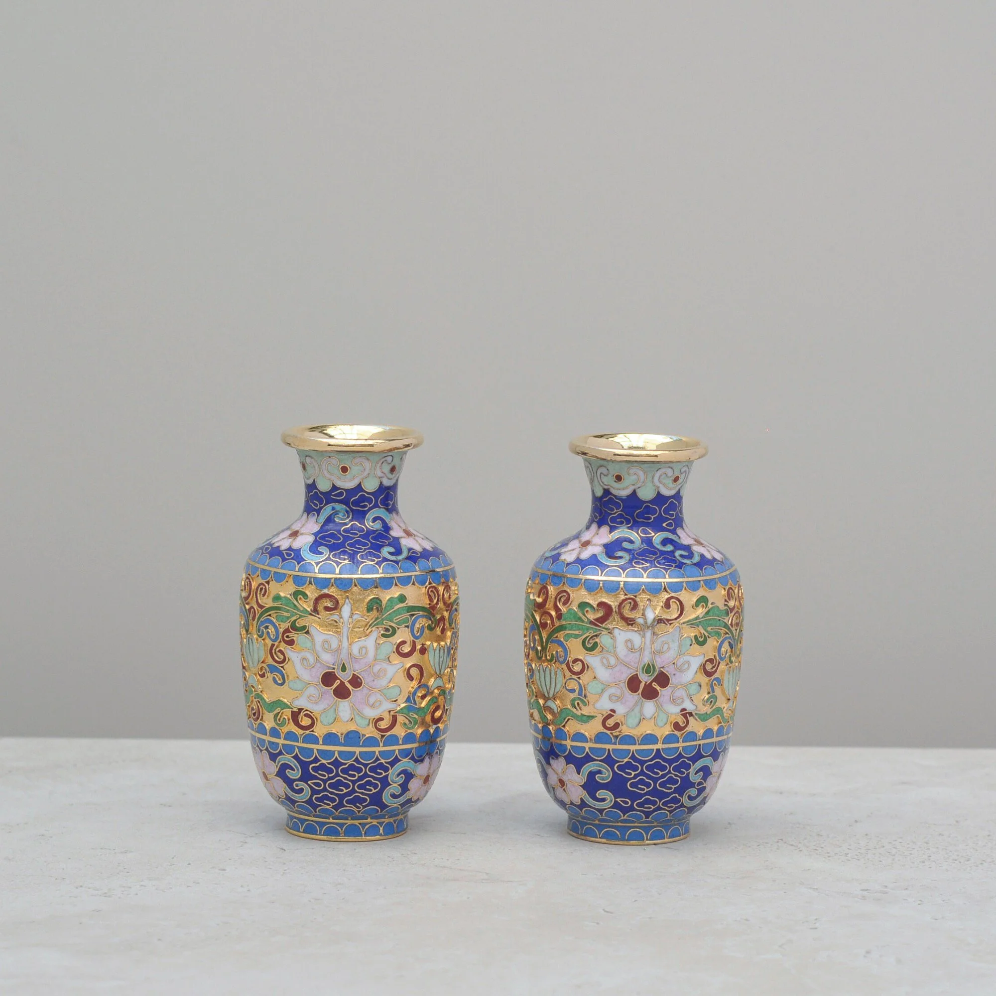Angled view of two mid-century Cloisonné vases featuring intricate blue, pink and gold enamel patterns with brass rims.