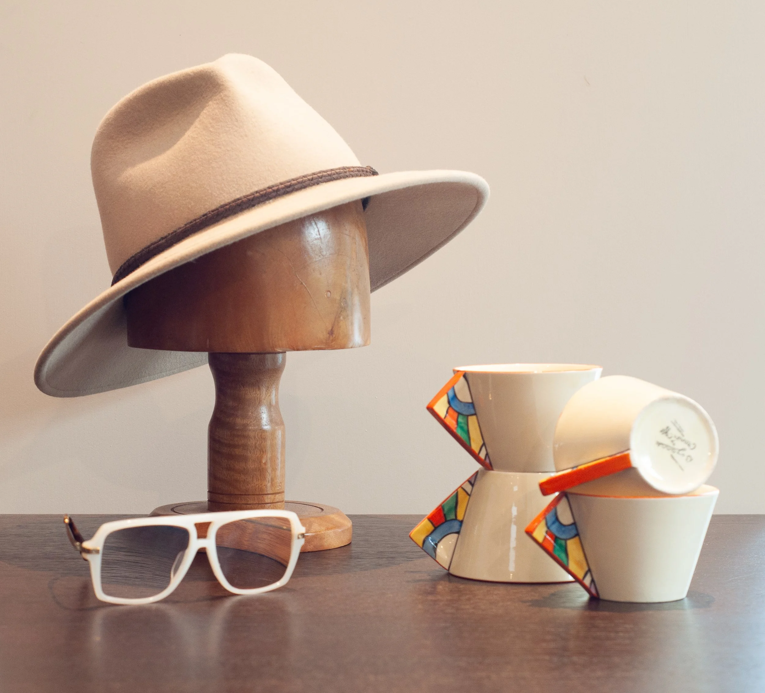 A beige wide-brimmed felt hat with a brown band on a wooden hat stand, white sunglasses with dark lenses on a dark wooden surface, and stacked white porcelain cups with colorful geometric patterns on their sides.