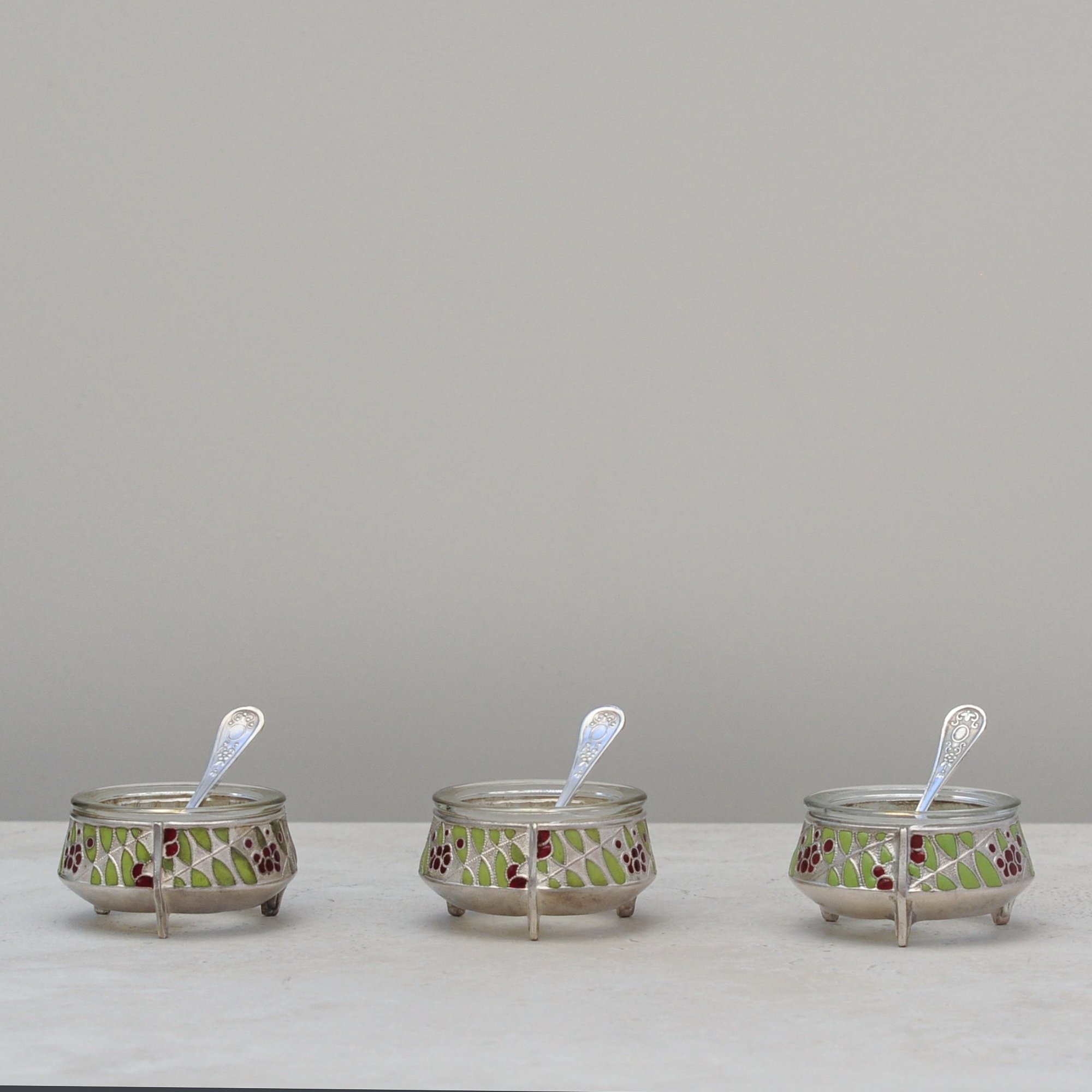 Three vintage silver-plated cloisonné salt cellars with enamelled birds and matching spoons, angled front view.
