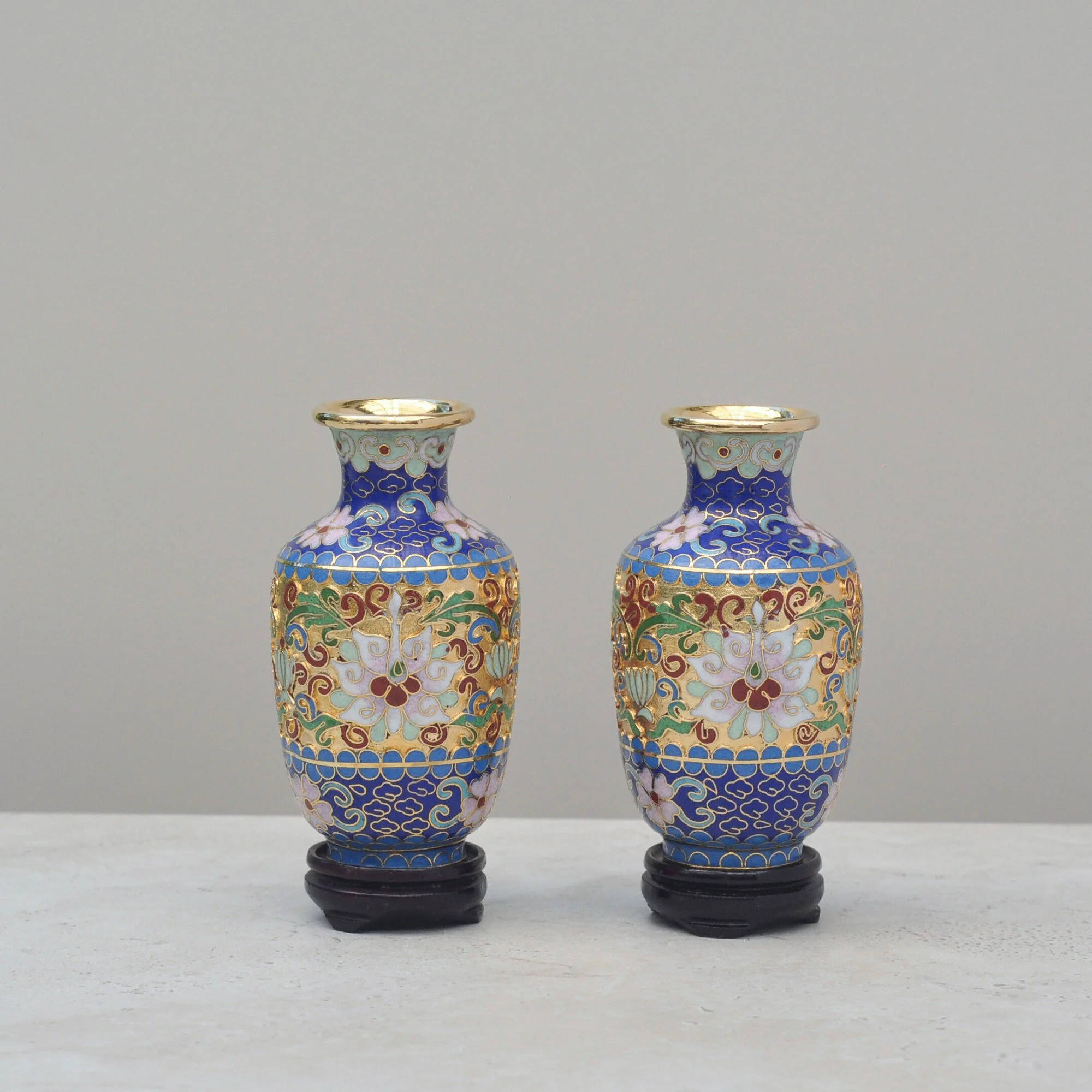 Styled scene featuring a pair of mid-century Cloisonné vases on wooden stands beside a vase