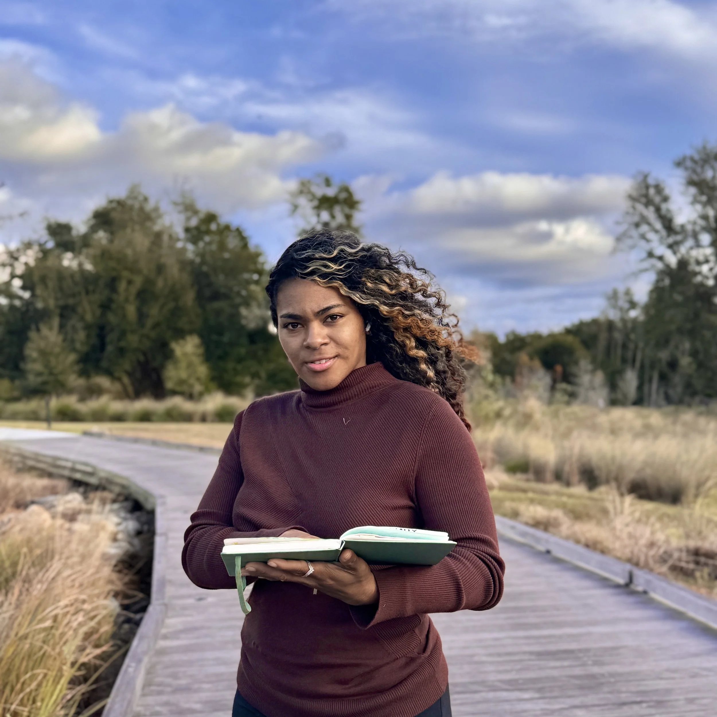 Young woman with curly hair holding an open notebook outdoors on a wooden walkway, with trees and a cloudy sky in the background.