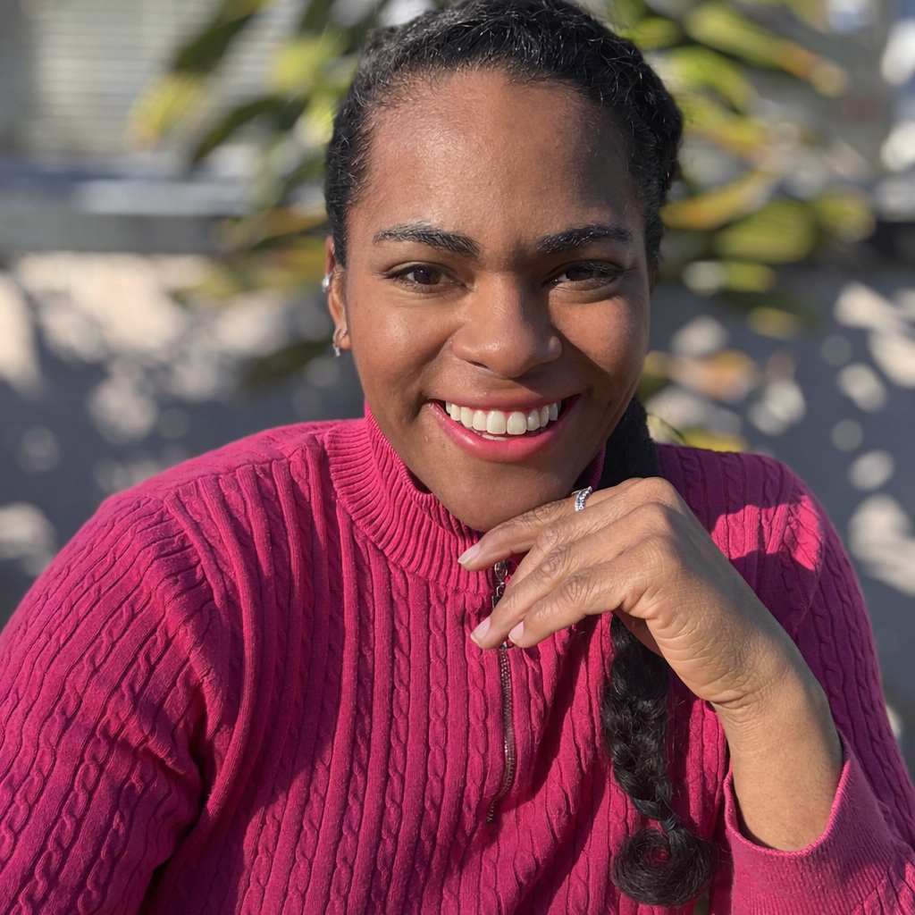 A smiling woman with dark braided hair wearing a pink sweater, resting her chin on her hand outdoors on a sunny day.