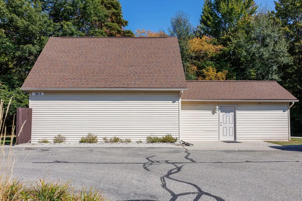 Single-story building with beige vinyl siding and a brown shingle roof, surrounded by trees with fall foliage, and a parking lot in the foreground.
