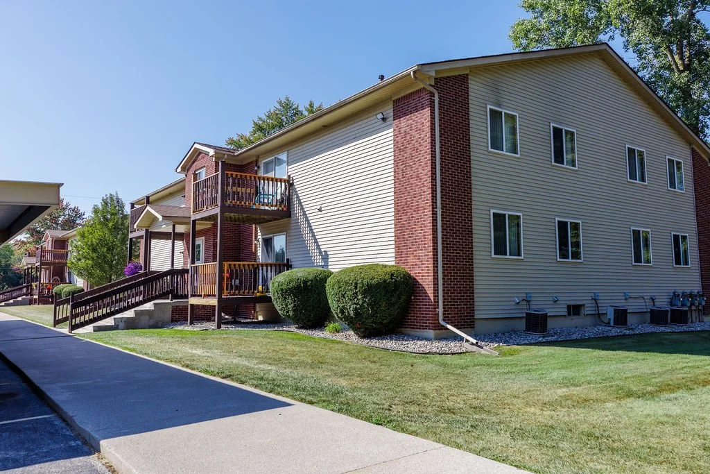 A multi-story apartment building with beige siding and red brick accents, featuring small balconies, surrounded by a well-maintained lawn, bushes, and trees under a clear blue sky.