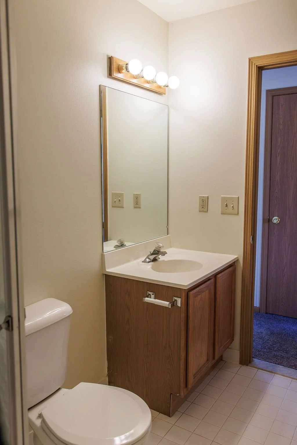Bathroom with a toilet, wooden vanity with a sink, mirror, and a row of four light bulbs above the mirror. There are electrical outlets and a door leading to another room.