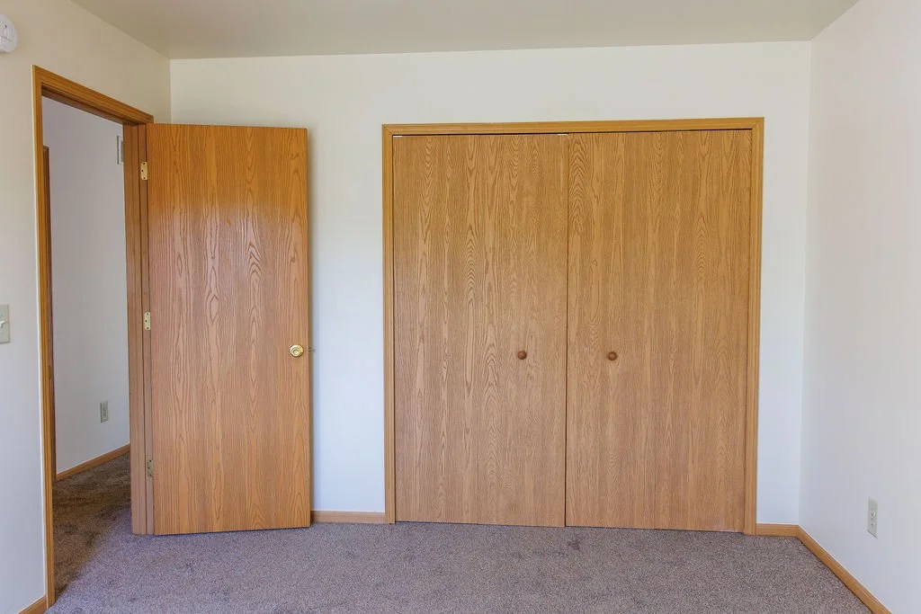 Empty room with a closet door and an open door leading to another room, tan carpet, white walls, and wooden trim.