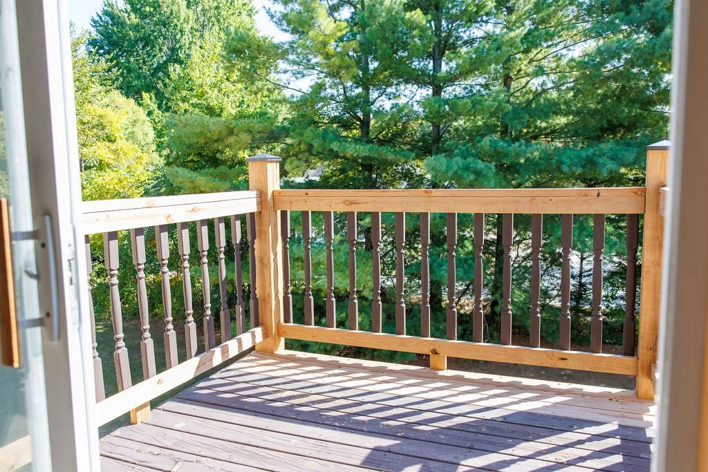View of a small wooden balcony with a rope railing, overlooking green trees under clear blue sky.