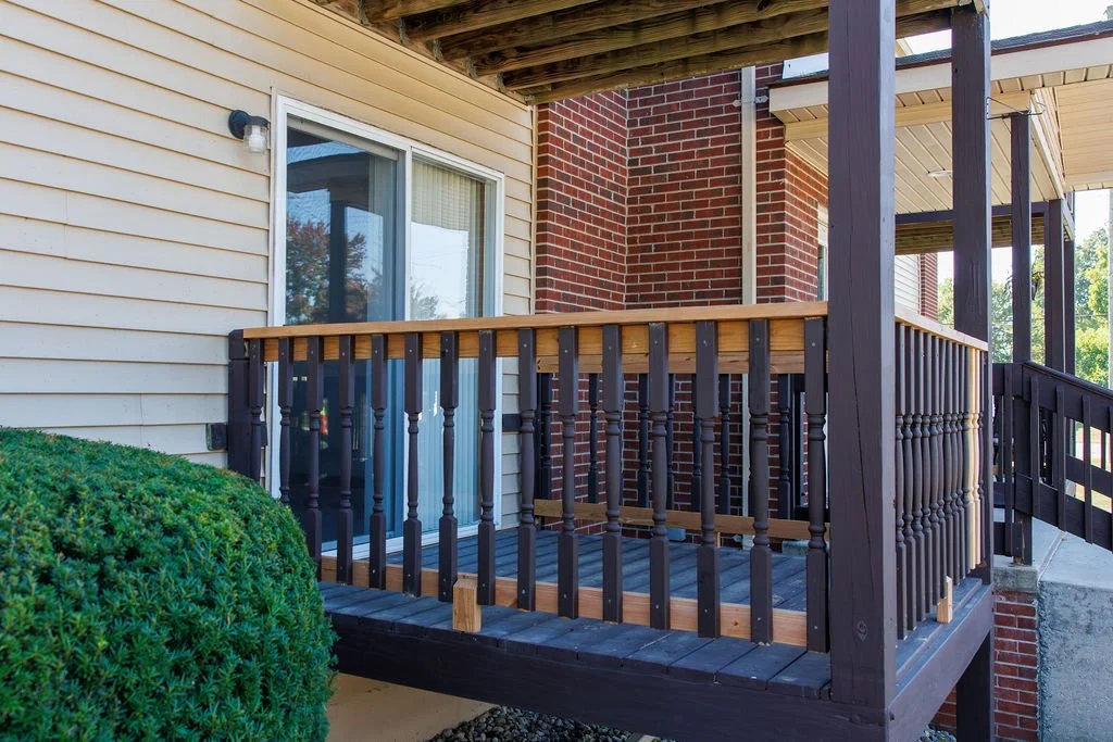 Image of a small wooden balcony attached to a house with beige siding, red brick wall, and a sliding glass door, surrounded by some greenery and a shrub.