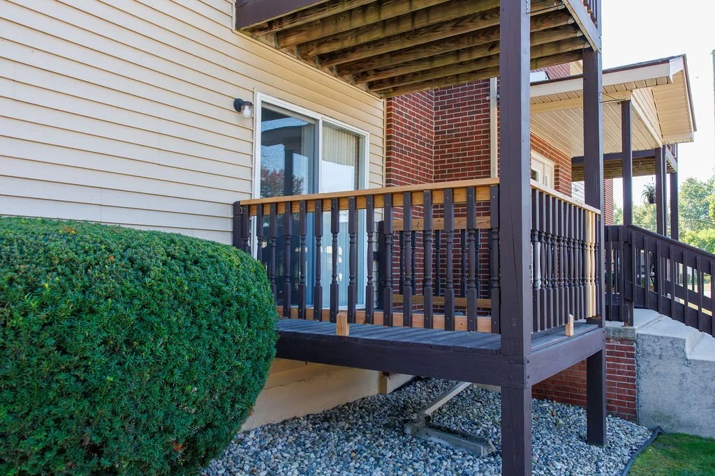 View of a small wooden balcony attached to a brick house, with a slide door leading inside. The balcony has dark brown railing and posts, with a wooden top rail. There is a large green bush in front of the house and a gravel ground beneath the balcon