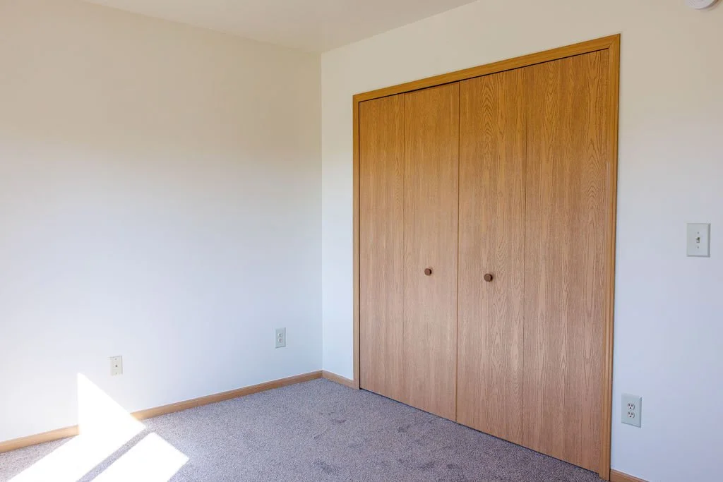 Empty room with white walls, beige carpet, wooden closet doors, and electrical outlets.