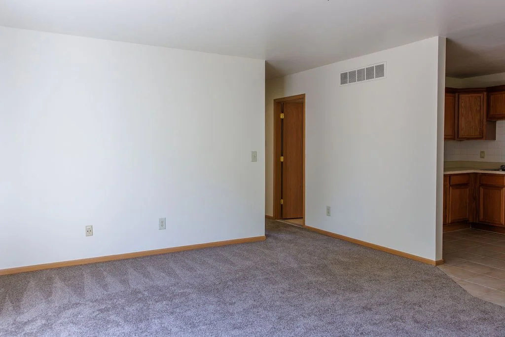 Empty living room with white walls, brown carpeting, and a doorway leading to a kitchen with wooden cabinets and tiled floor.