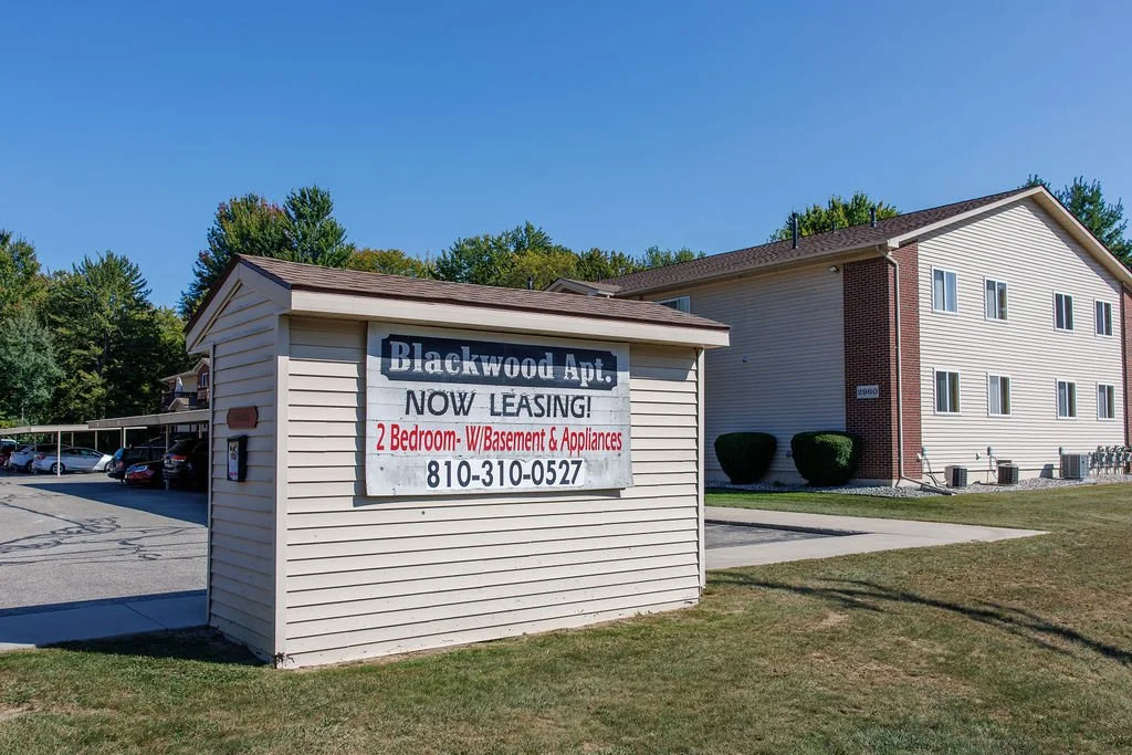Apartment complex with a sign advertising 2-bedroom apartments for lease. The sign on the small building reads 'Blackwood Apt. Now Leasing! 2 Bedroom- W/Basement & Appliances, 810-310-0527.' There are parking spaces and apartment buildings in the bac