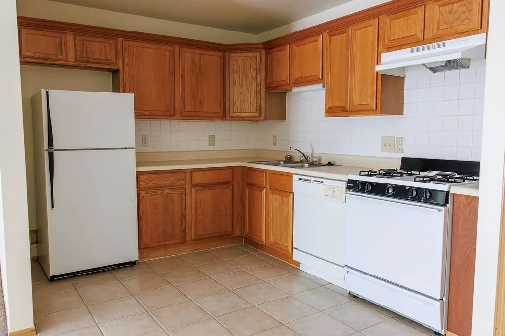 Kitchen with wooden cabinets, white appliances including refrigerator, dishwasher, and stove, beige countertops, white tiled backsplash, and tiled floor.