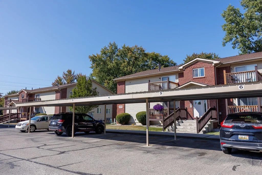 Apartment complex with numbered parking carport, cars parked underneath, and residential units with stairs and balconies.