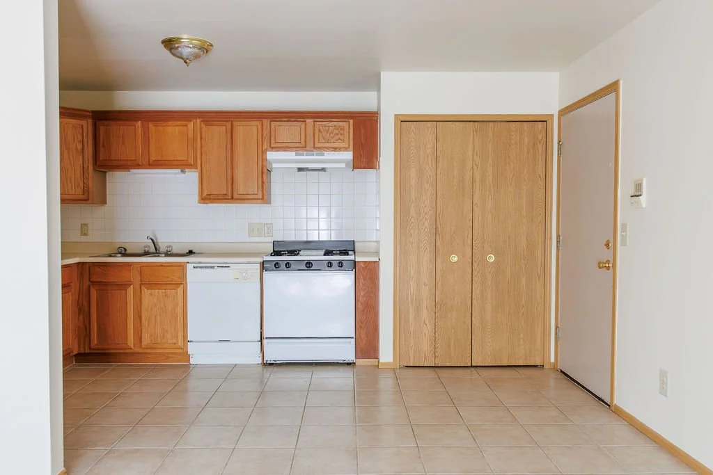 Empty kitchen with wooden cabinets, white appliances, tiled floor, a closet with wooden doors, and a front door with a doorbell and thermostat.