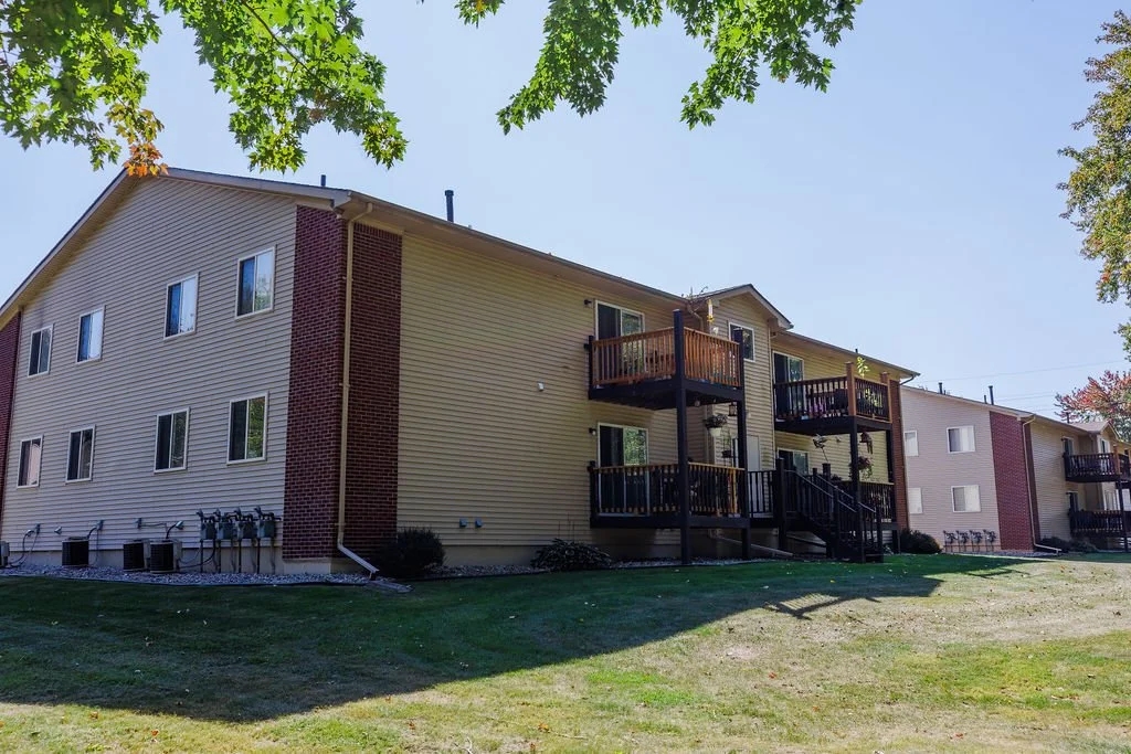 Apartment complex with multiple units, yellow siding, red brick accents, and small outdoor decks, surrounded by a grassy lawn and trees.