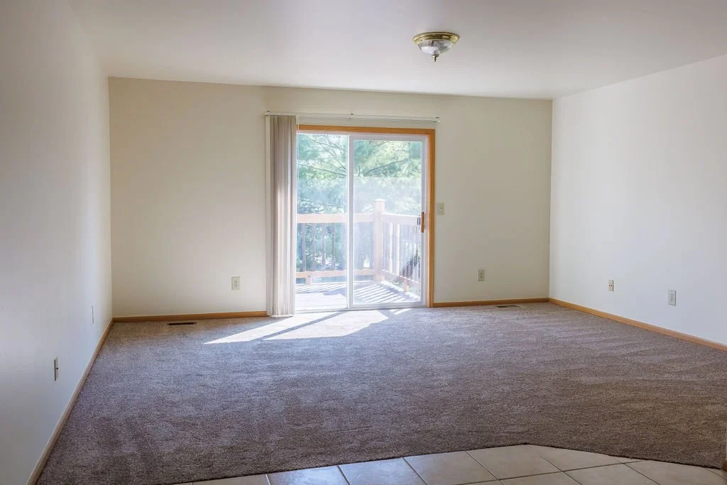 Empty living room with sliding glass door leading to a balcony, beige carpet, white walls, and ceiling light fixture.