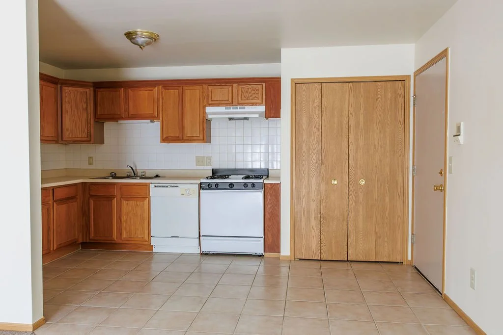 Empty kitchen with wooden cabinets, white appliances, tile flooring, and a closet door.