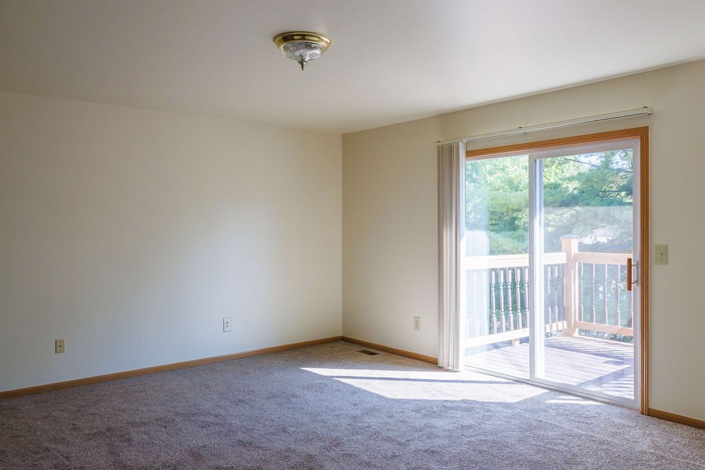 Empty living room with sliding glass door leading to a wooden deck, sunlight streaming inside, beige carpet, off-white walls, and a ceiling light fixture.