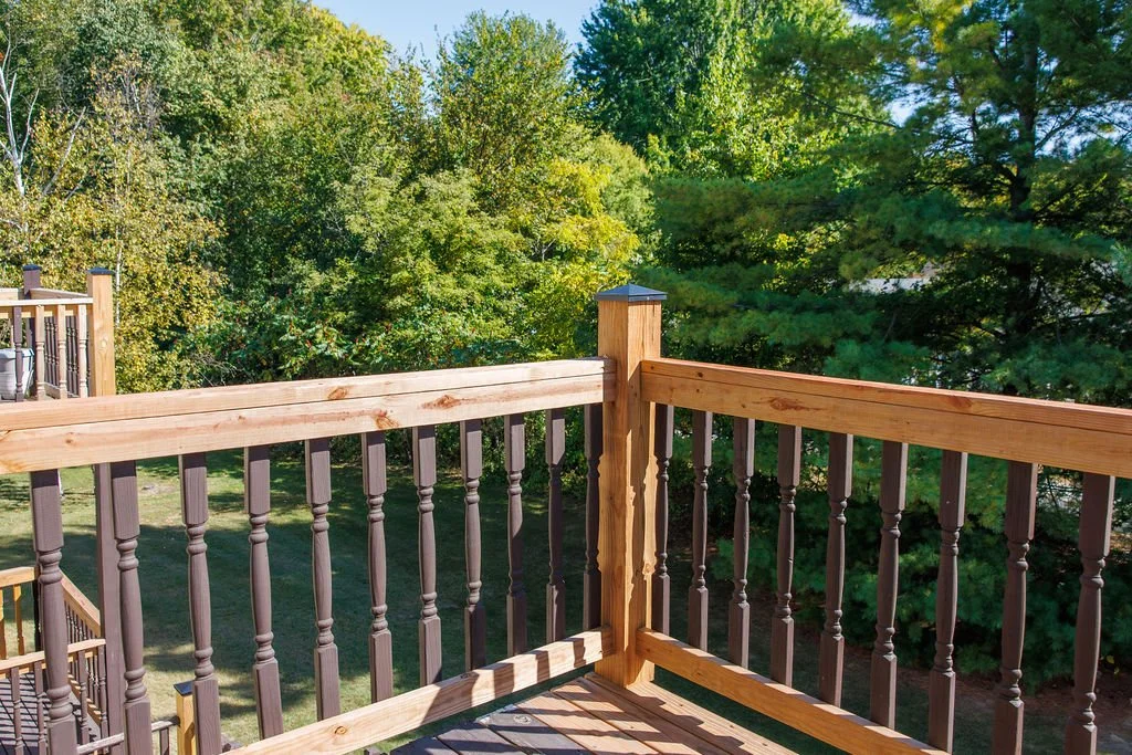 Wooden balcony or deck with railing overlooking a green yard with trees, under clear blue sky.