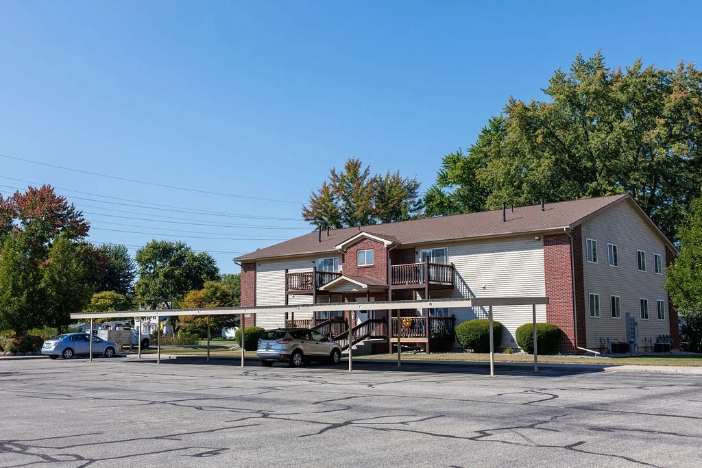 Two-story residential building with a parking lot in front, trees in the background, and a clear blue sky.