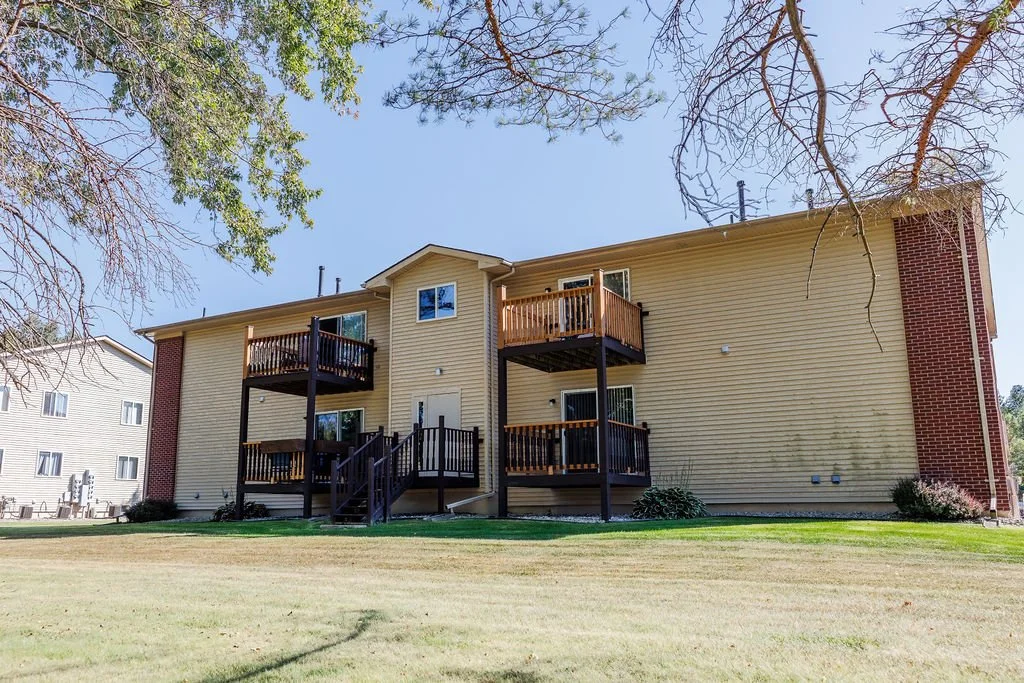Apartment building with beige siding, red brick accents, and small wooden balconies. Green grass and trees in the foreground, clear blue sky overhead.