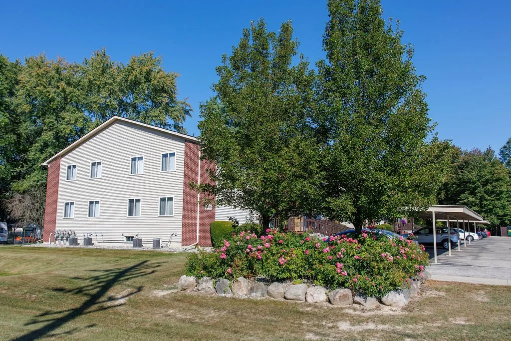 A residential apartment building with multiple windows, a sloped roof, and beige siding with red brick accents, next to a large green tree with pink flowering bushes at its base, and a parking lot with several cars on the right.