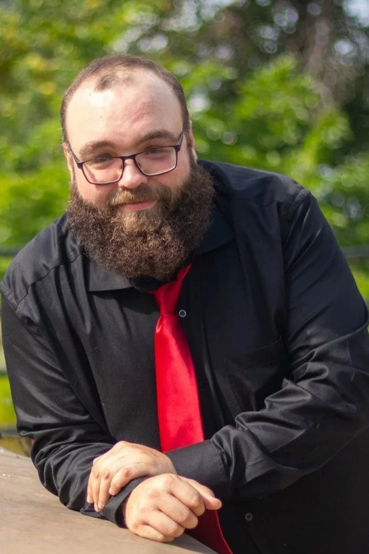 A man with glasses and a beard leaning on a wooden surface outdoors, wearing a black shirt and a red tie, with green blurred foliage in the background.