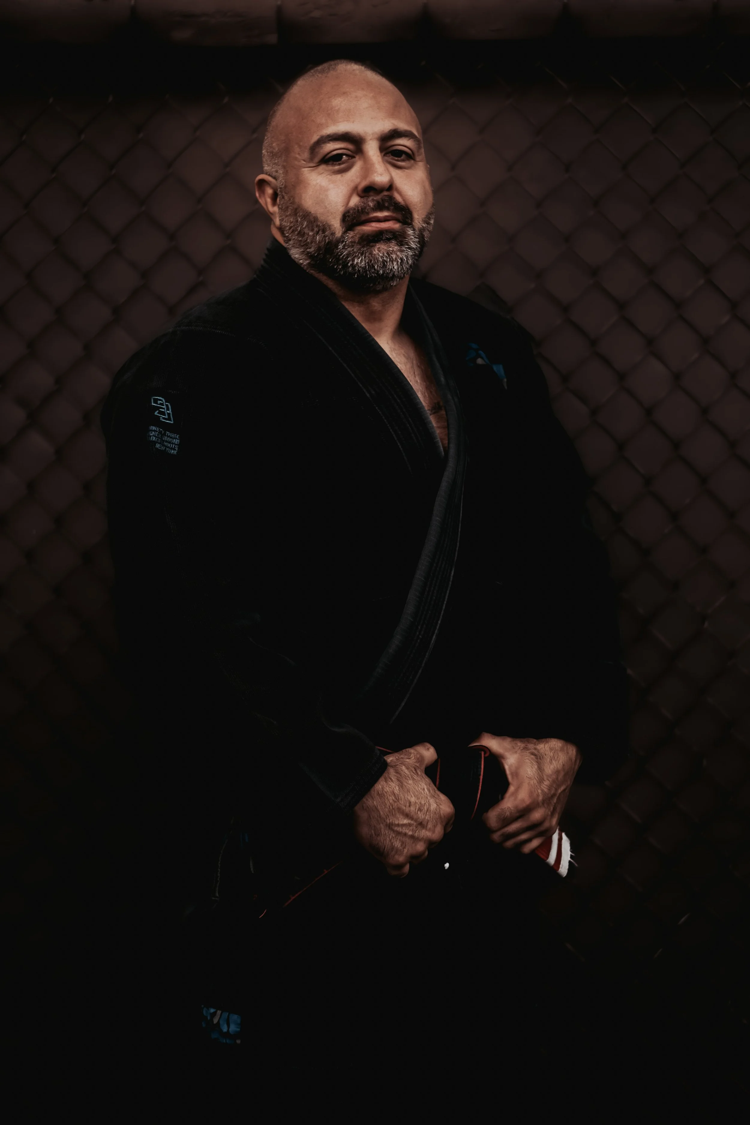 Martial artist in black gi standing in front of a chain-link fence.