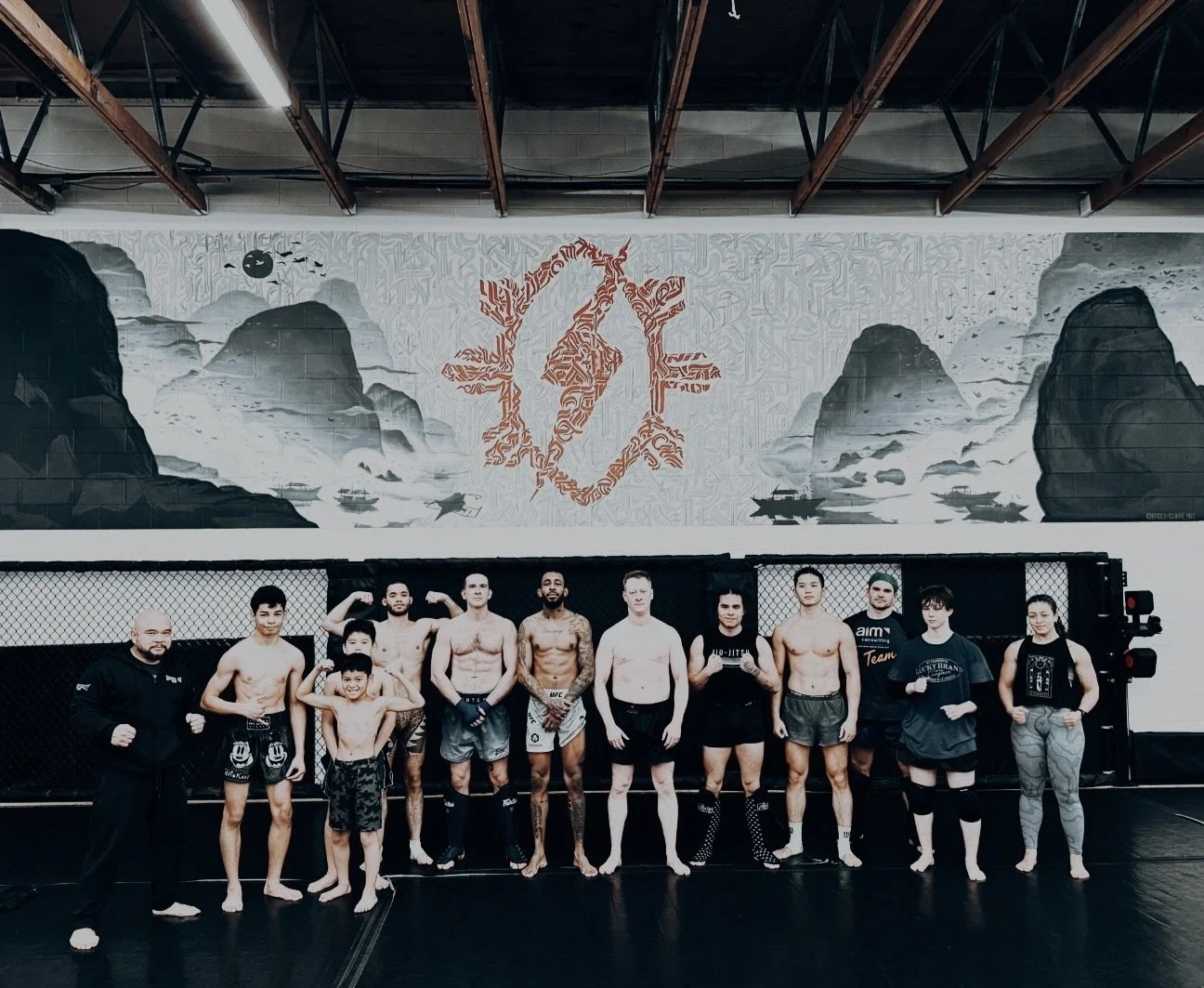 Group of people in a gym wearing martial arts attire, standing together on a training mat with a mural on the wall behind them.