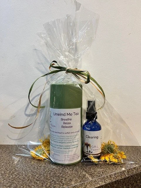 A gift set containing a green bottle labeled 'Unwind Me Tea' and a smaller blue spray bottle labeled 'Clearing', wrapped together in clear plastic with a decorative ribbon and dried flowers.