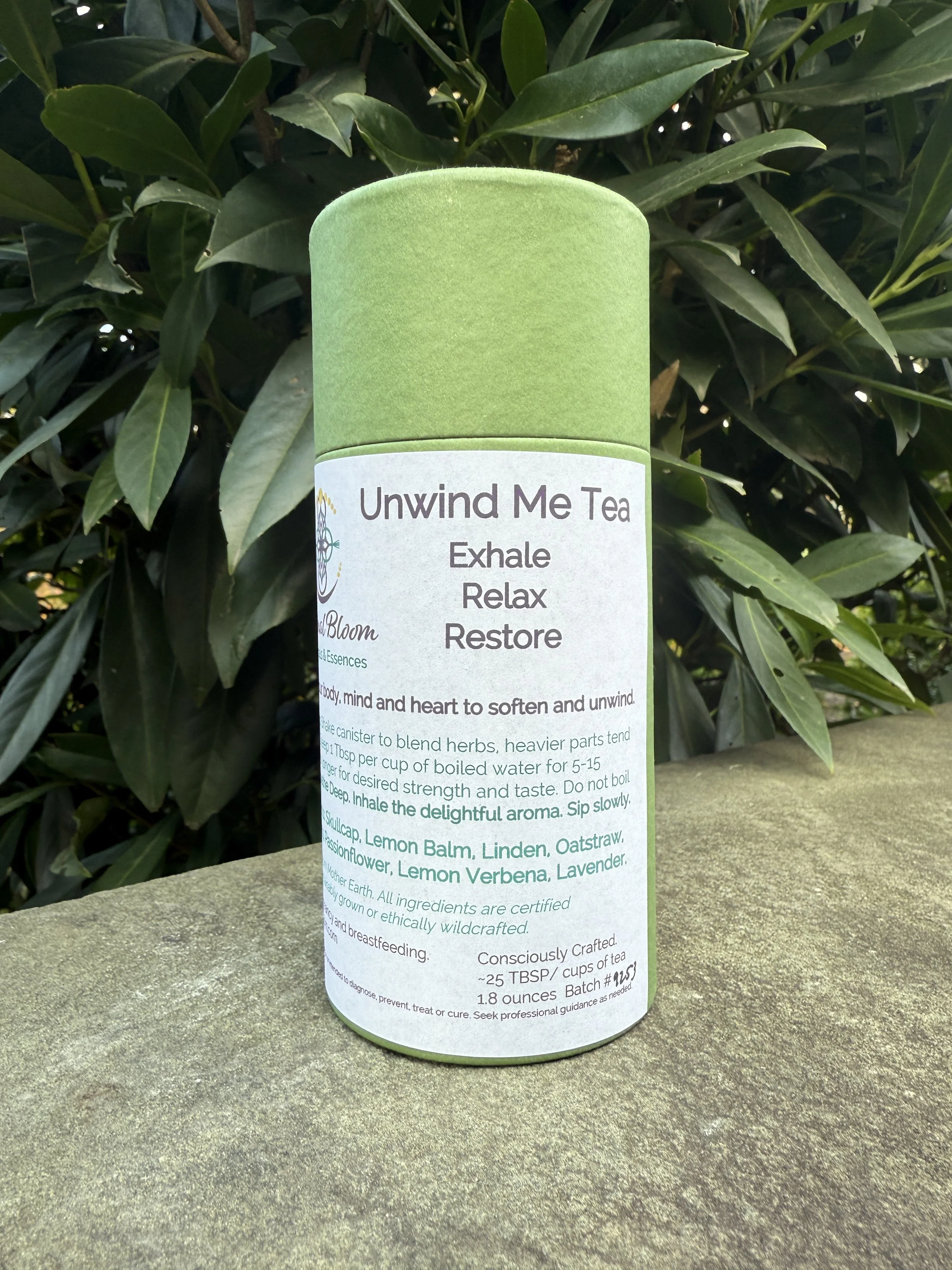 A cylindrical green tea container labeled "Unwind Me Tea" with instructions and ingredients, placed on a stone surface with green foliage in the background.
