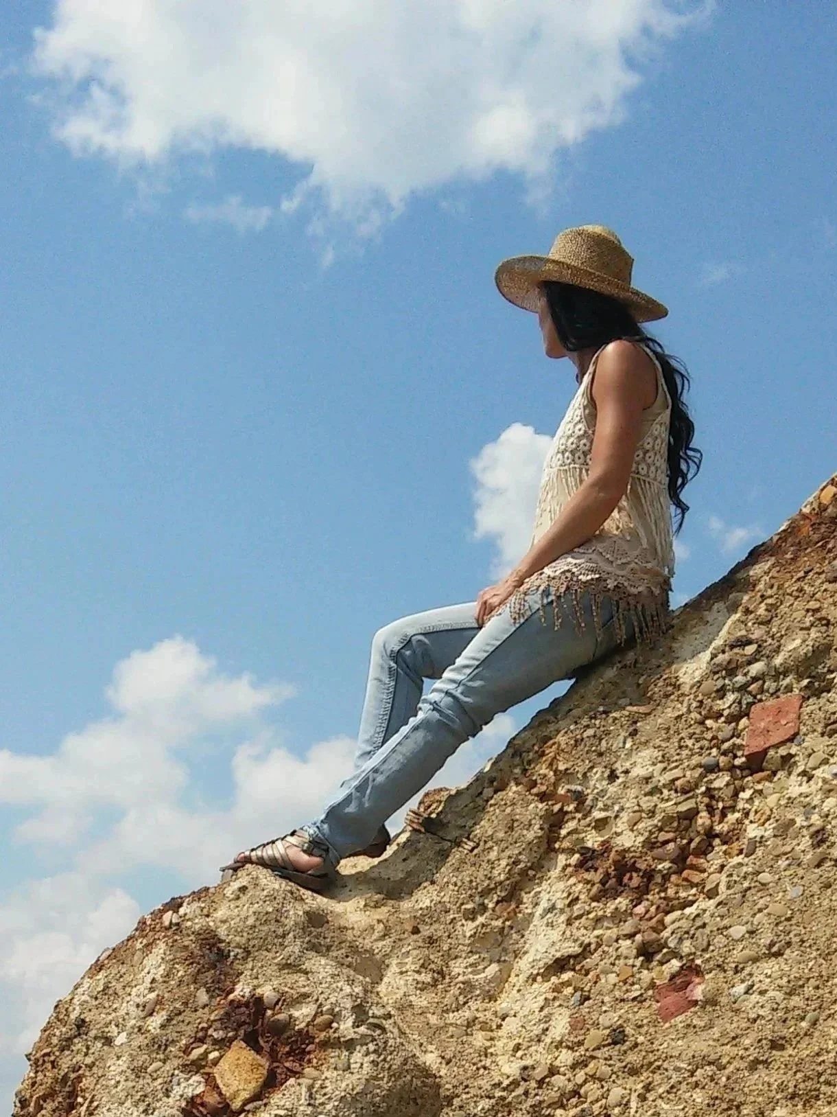 A woman with long dark hair sitting on a rocky hill, wearing a wide-brimmed straw hat, a beige sleeveless top, light wash jeans, and sandals, looking into the distance with a blue sky and scattered clouds in the background.