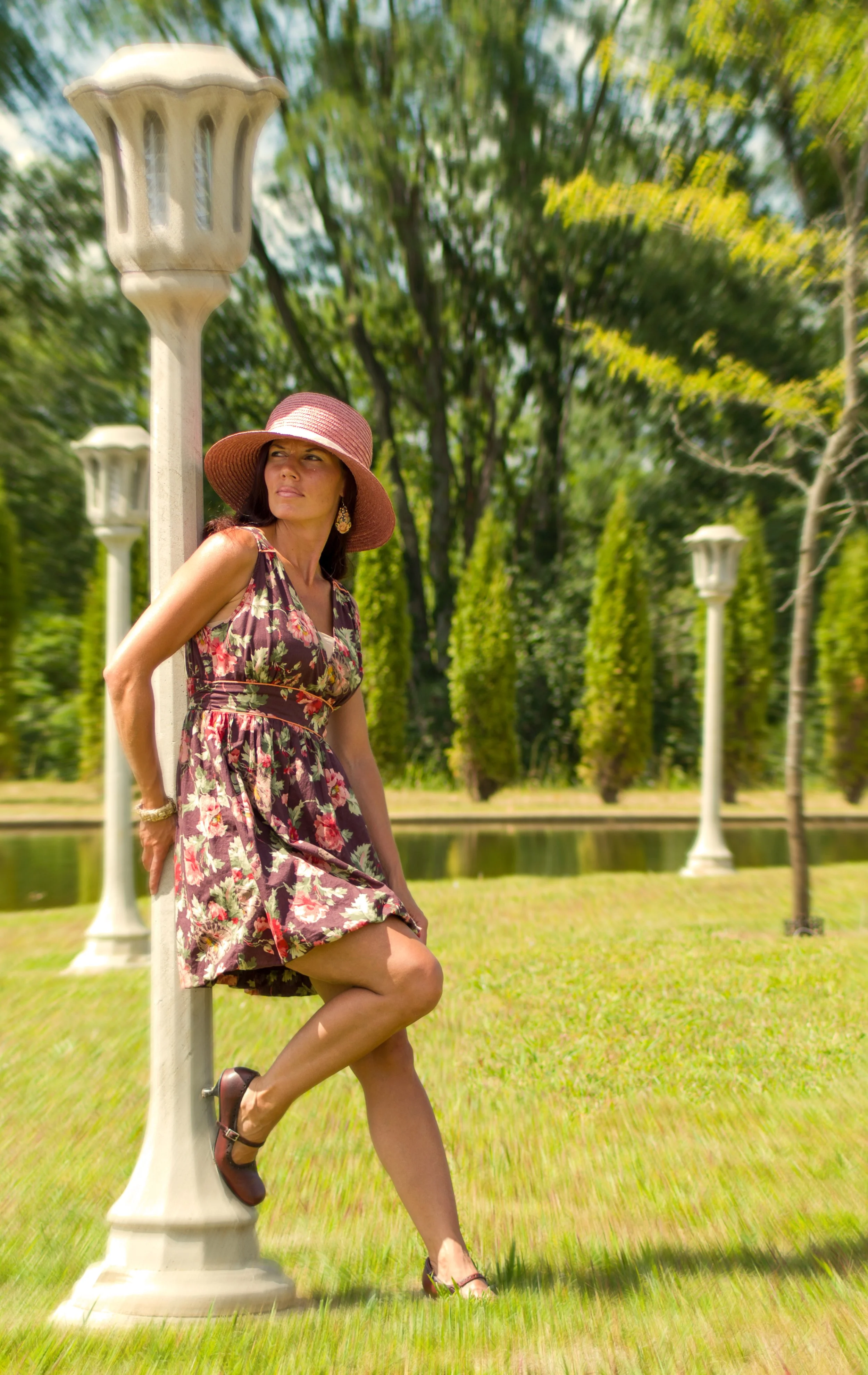 A woman wearing a floral dress and a wide-brimmed pink hat stands outdoors near a vintage lamp post in a park with green trees and a pond in the background.