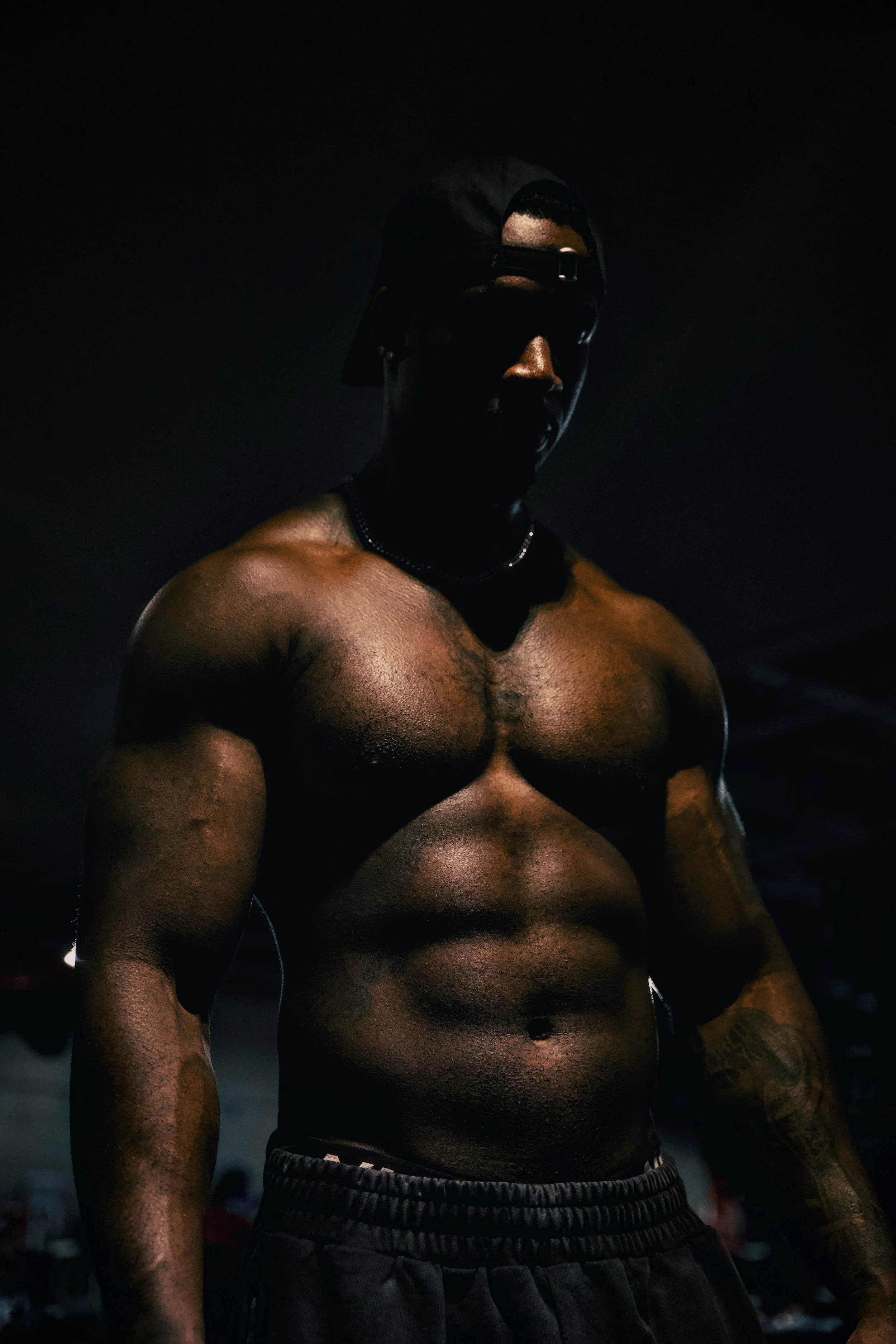 A shirtless man with a muscular build wearing a backward cap and sunglasses, standing in a dark environment with his face partially obscured by shadows.
