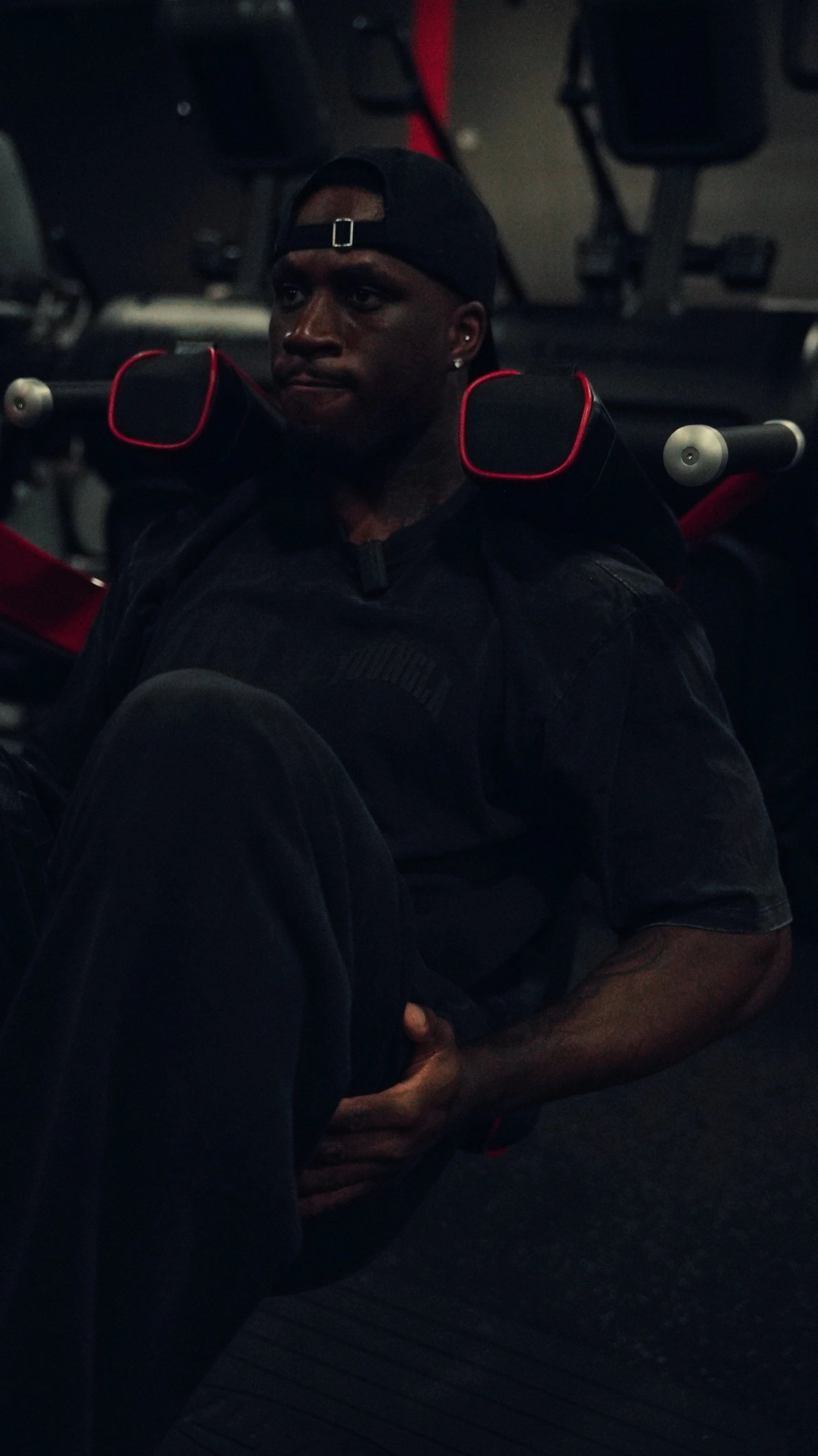 A man in a black cap and black clothing sitting on the floor with his back against gym equipment, holding his knee with one hand, in a dimly lit gym.