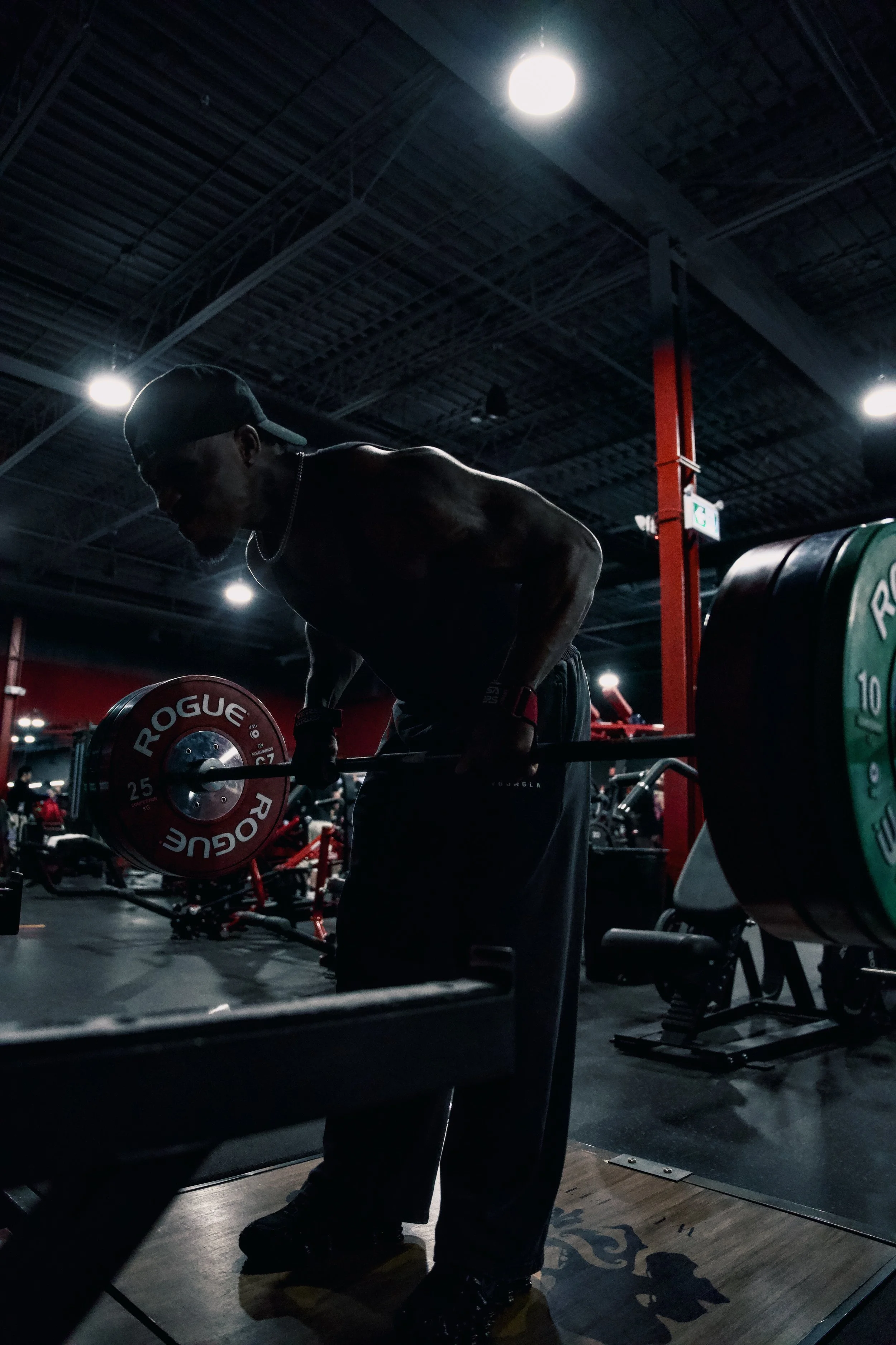 A man lifting weights at a gym in a dark, dimly lit environment with red and black equipment in the background.